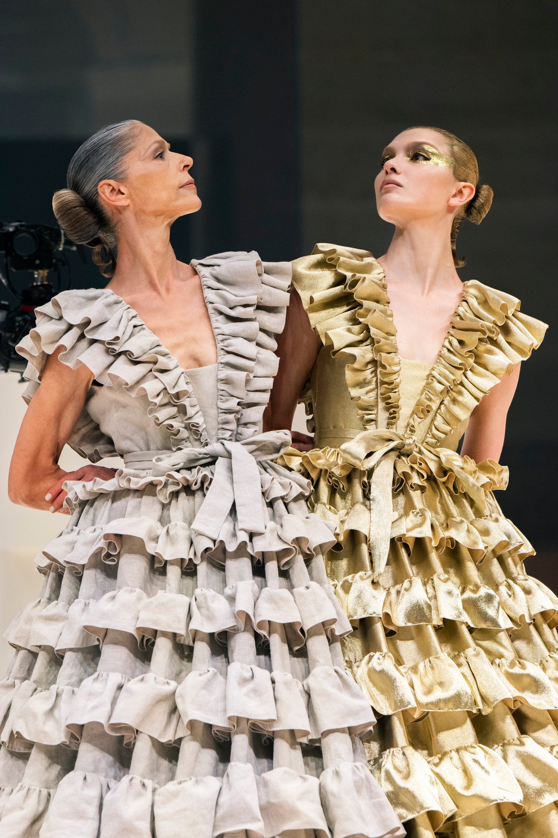Two models wearing dramatic, heavily ruffled evening gowns. The model on the left wears a deep V-neck gown in a light warm gray or off-white fabric with cascading ruffles.