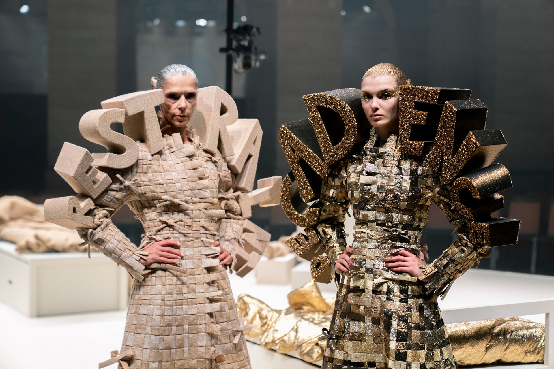 Two models walk a runway wearing highly structured, sculptural dresses made of stacked rectangular panels. The dress on the left is light beige/tan, and the dress on the right is metallic gold and black.