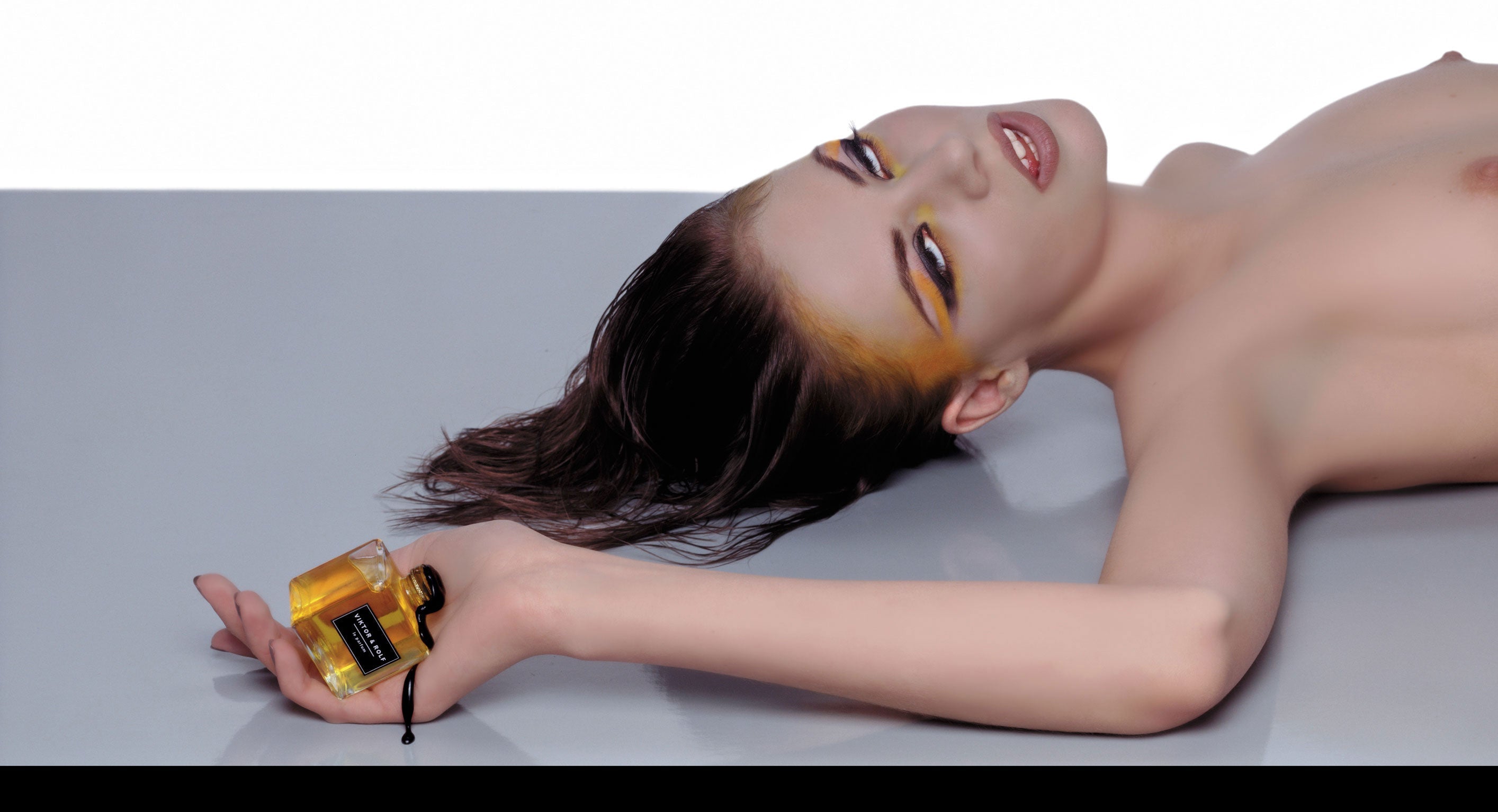 A model lies on a gray surface holding a Viktor & Rolf Le Parfum bottle. The model has dramatic yellow and orange eye makeup, and the image includes a black banner at the bottom with the text 'VIKTOR & ROLF LE PARFUM'.