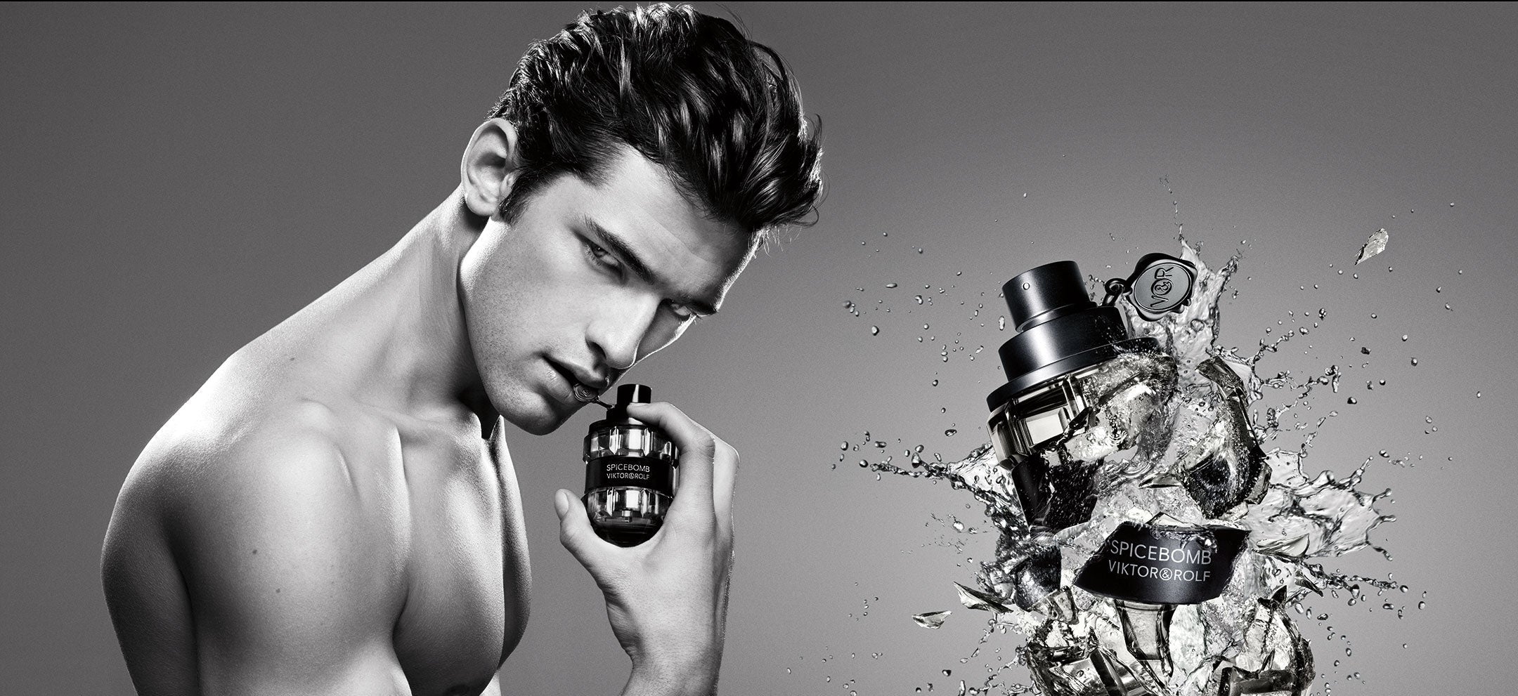 A shirtless male model holds a small cylindrical perfume bottle against his face, while a larger shattered glass perfume bottle with liquid splashing appears to the right. The image is monochrome and clearly advertising a fragrance.