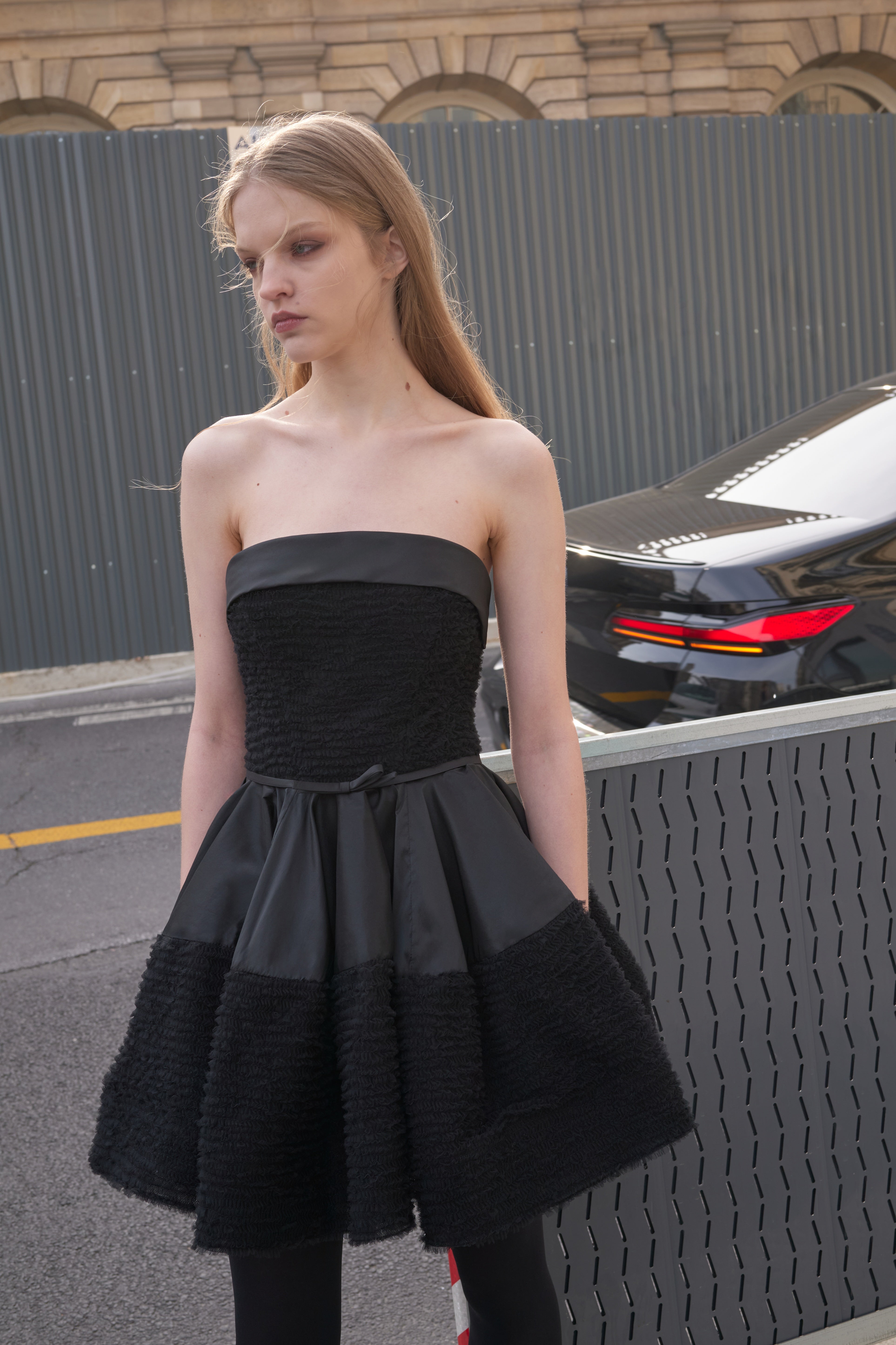 Model wearing a black strapless dress, standing next to a road.
