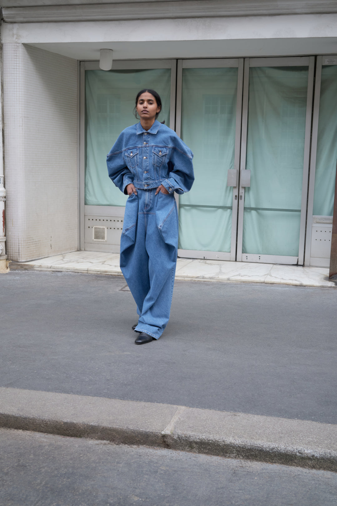Person wearing a blue denim outfit standing on a street with a building in the background.