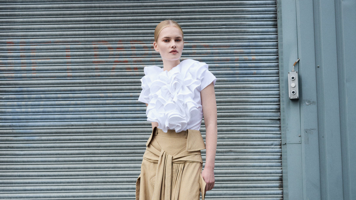Woman wearing a white ruffled top and beige pants standing in front of a metal shutter.