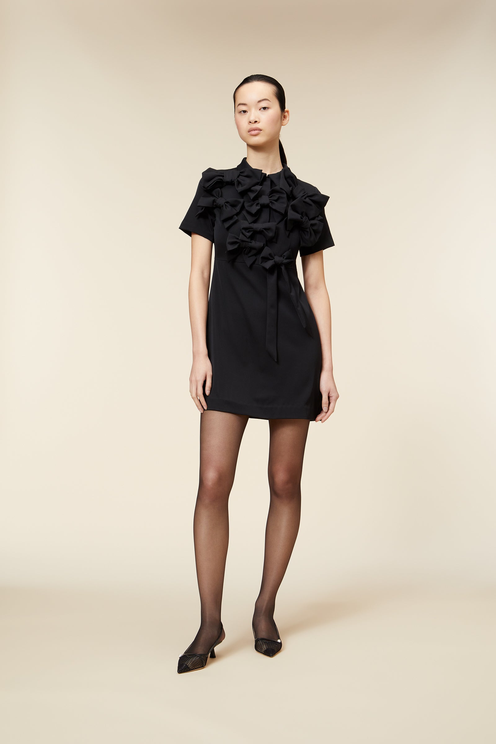 Model wearing elegant short black dress with large fabric bows, set against a neutral background.