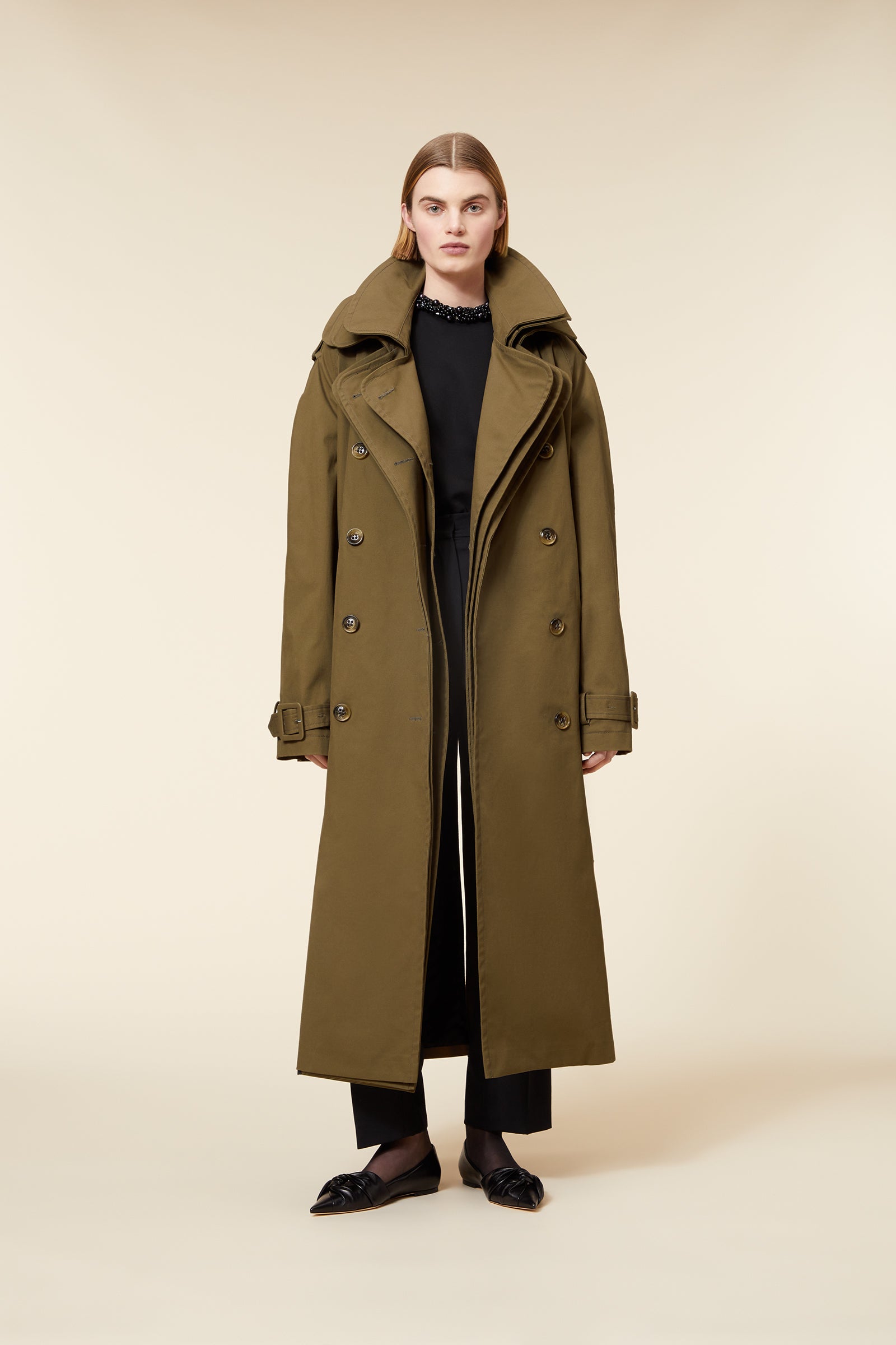 Olive green long trench coat featuring wide lapels, double-breasted buttons, and a belted waist, paired with black pants.
