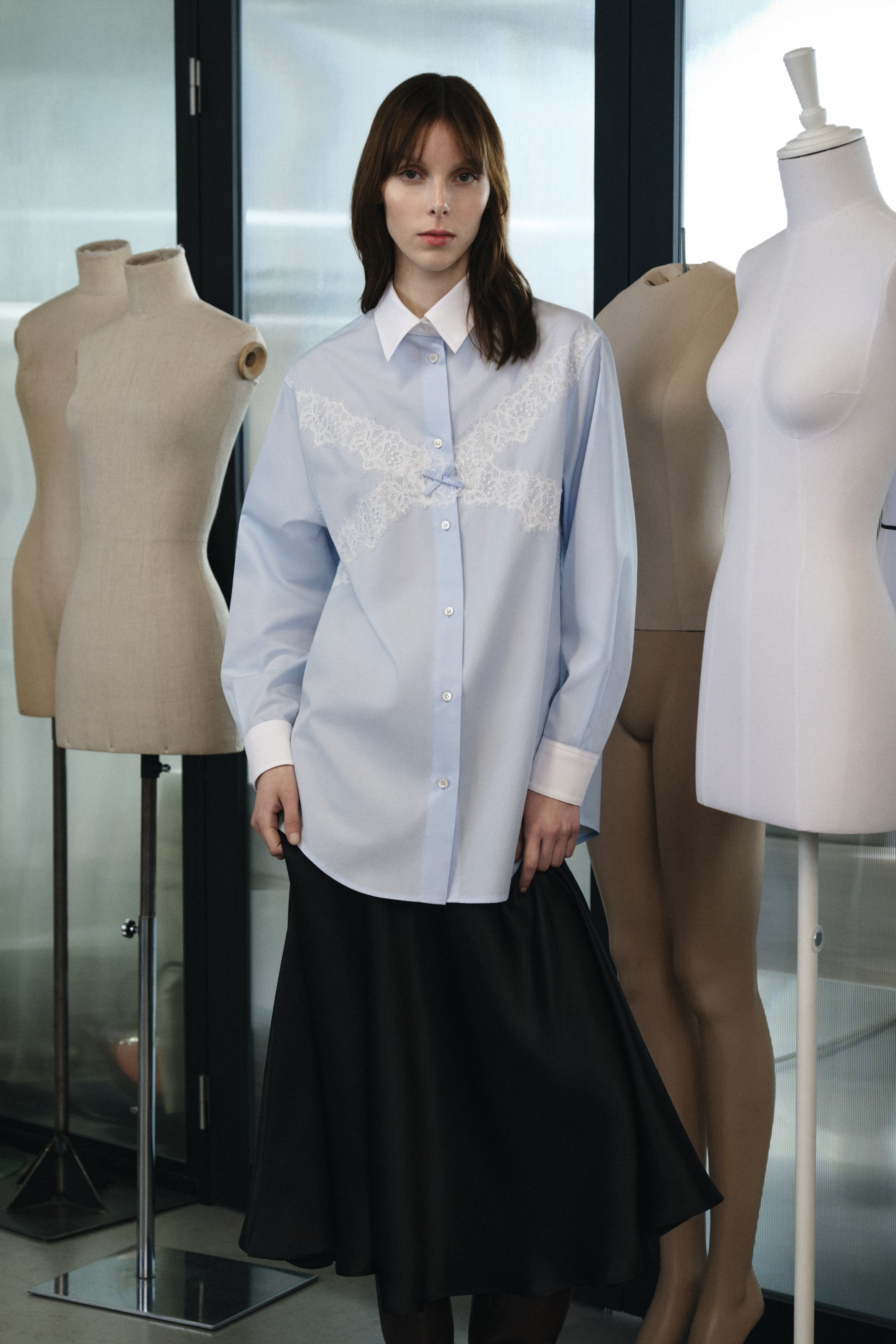 Woman wearing a light blue blouse with lace details in a fashion setting with mannequins.