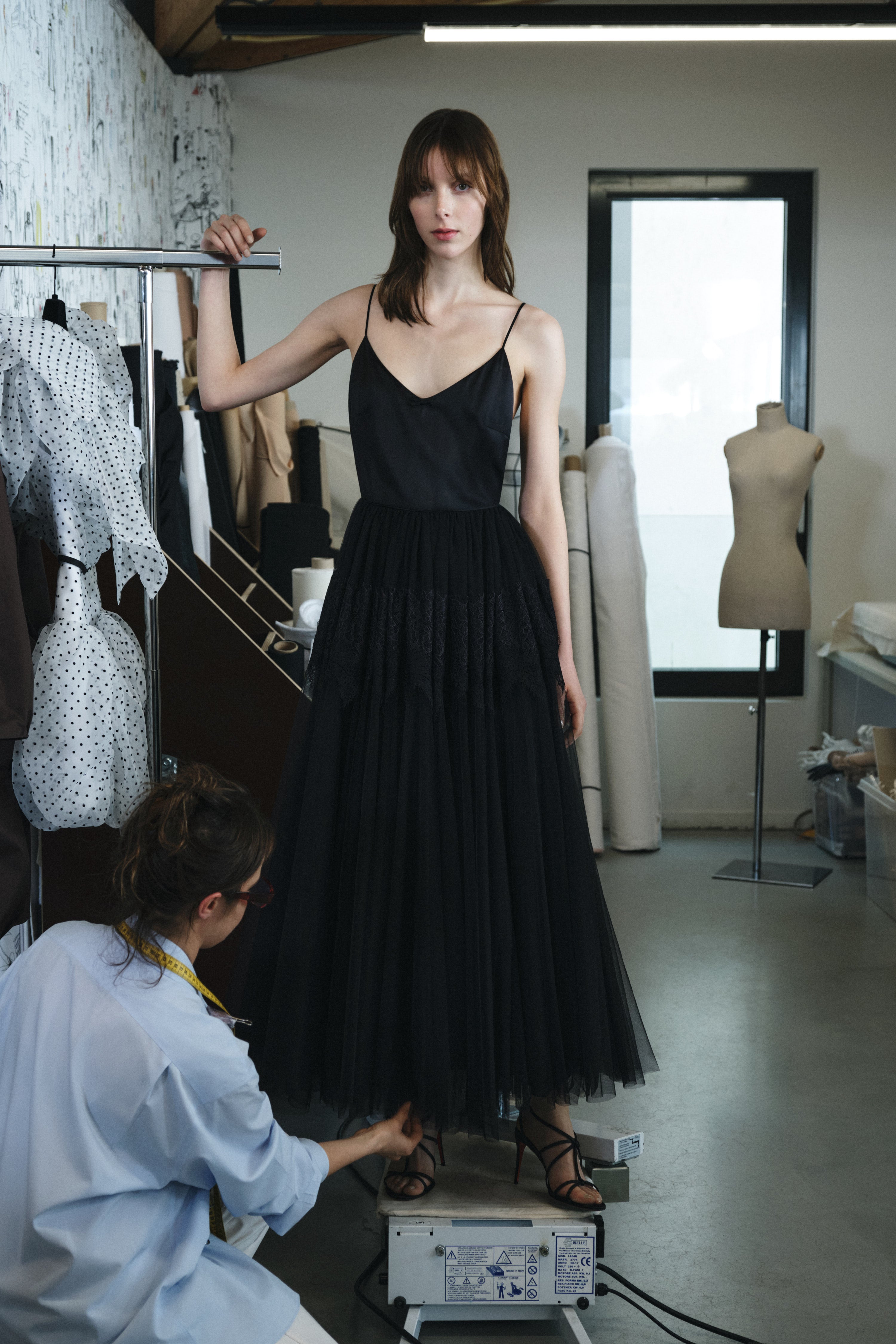 Model wearing a black dress in a fashion studio with another person adjusting it.