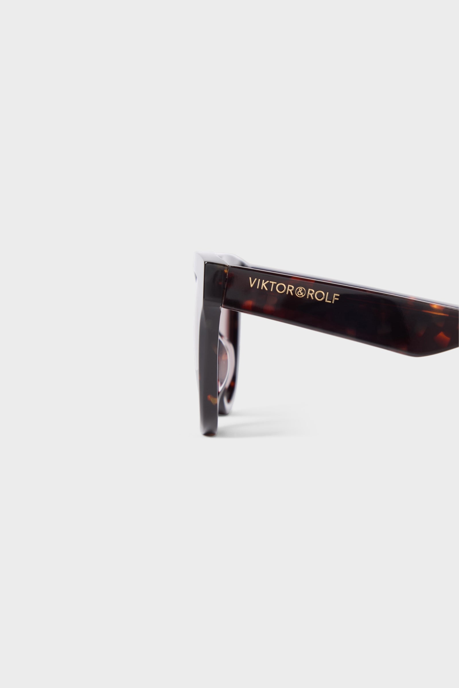 Tortoiseshell sunglasses arm featuring a sleek design with the brand logo in gold lettering against a light solid background.