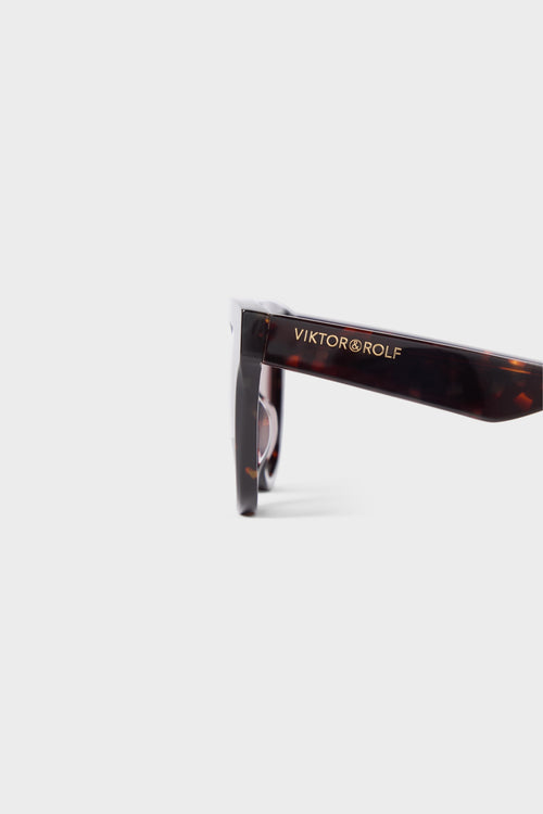 Tortoiseshell sunglasses arm featuring a sleek design with the brand logo in gold lettering against a light solid background.