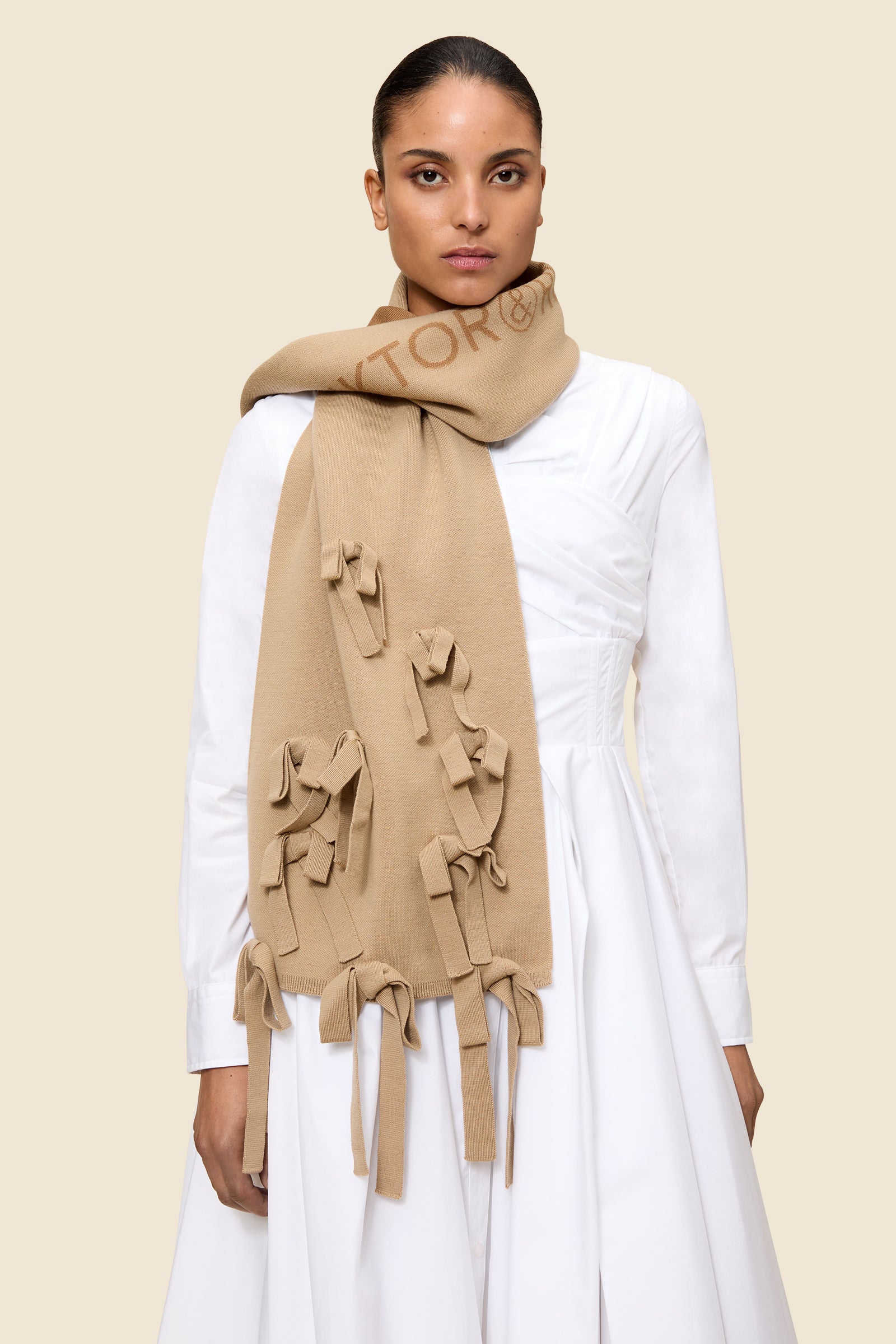 Elegant beige scarf with intricate bow details draped over a stylishly pleated white dress, creating a chic ensemble.