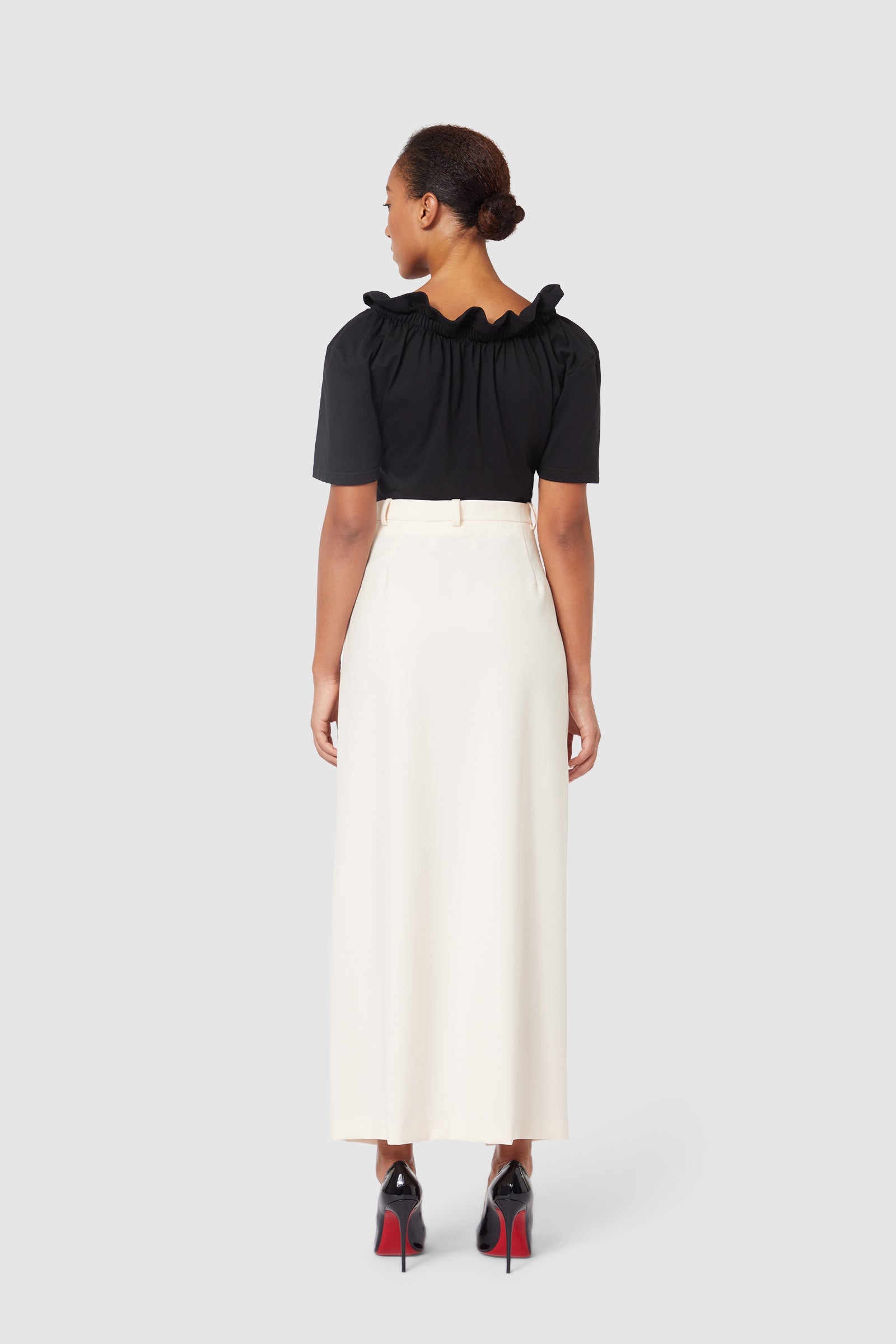A woman in a black ruffled top and cream high-waisted skirt, paired with black stiletto heels with red soles.