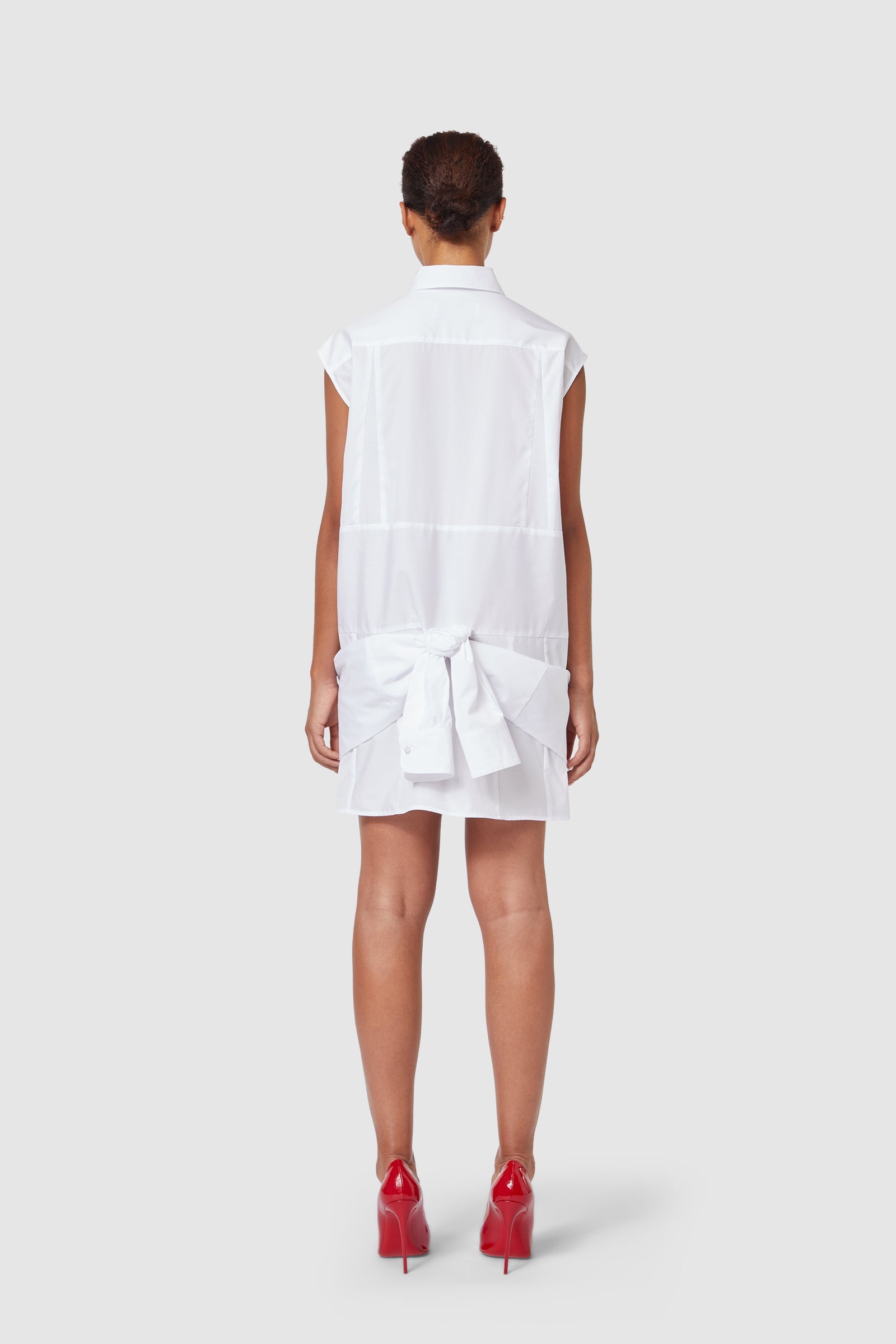 Sleeveless white shirtdress with back bow detailing, paired with red high heels, showcasing minimalist elegance.