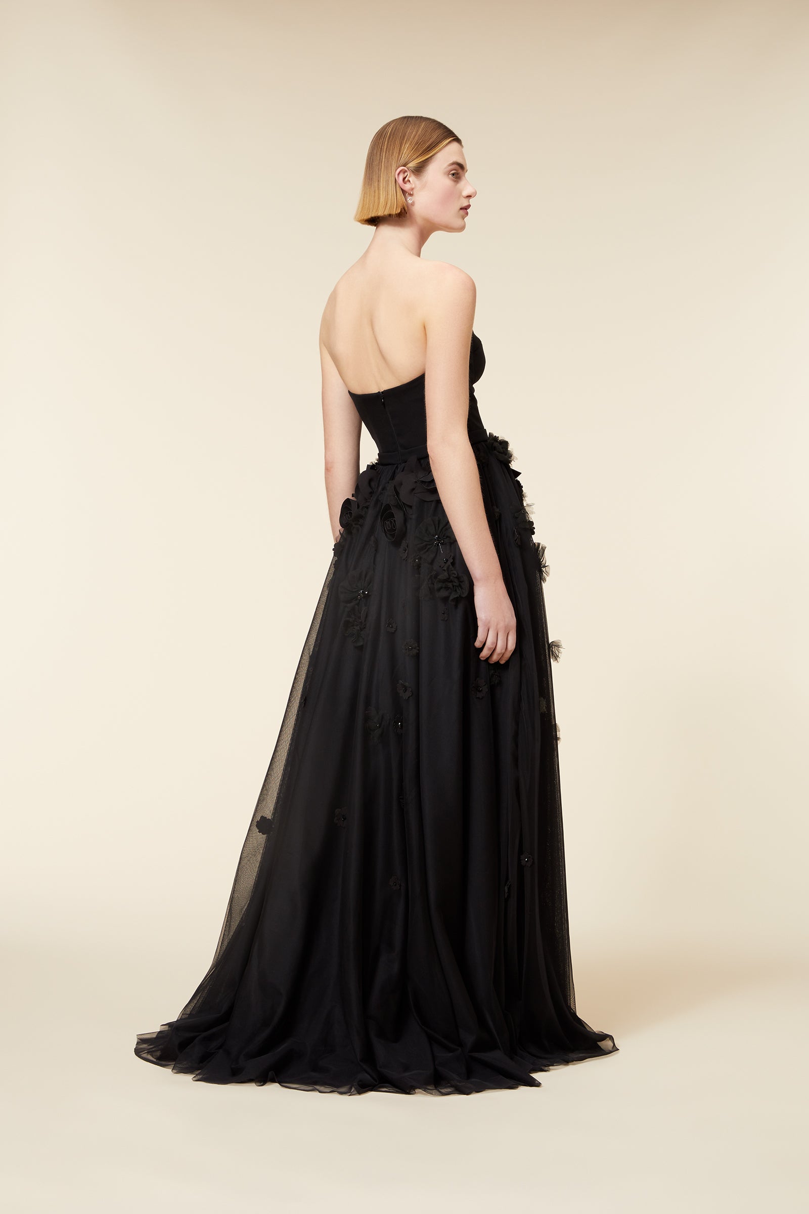Elegant black sleeveless dress with intricate floral appliqué detailing, worn by a woman in a minimalist setting.