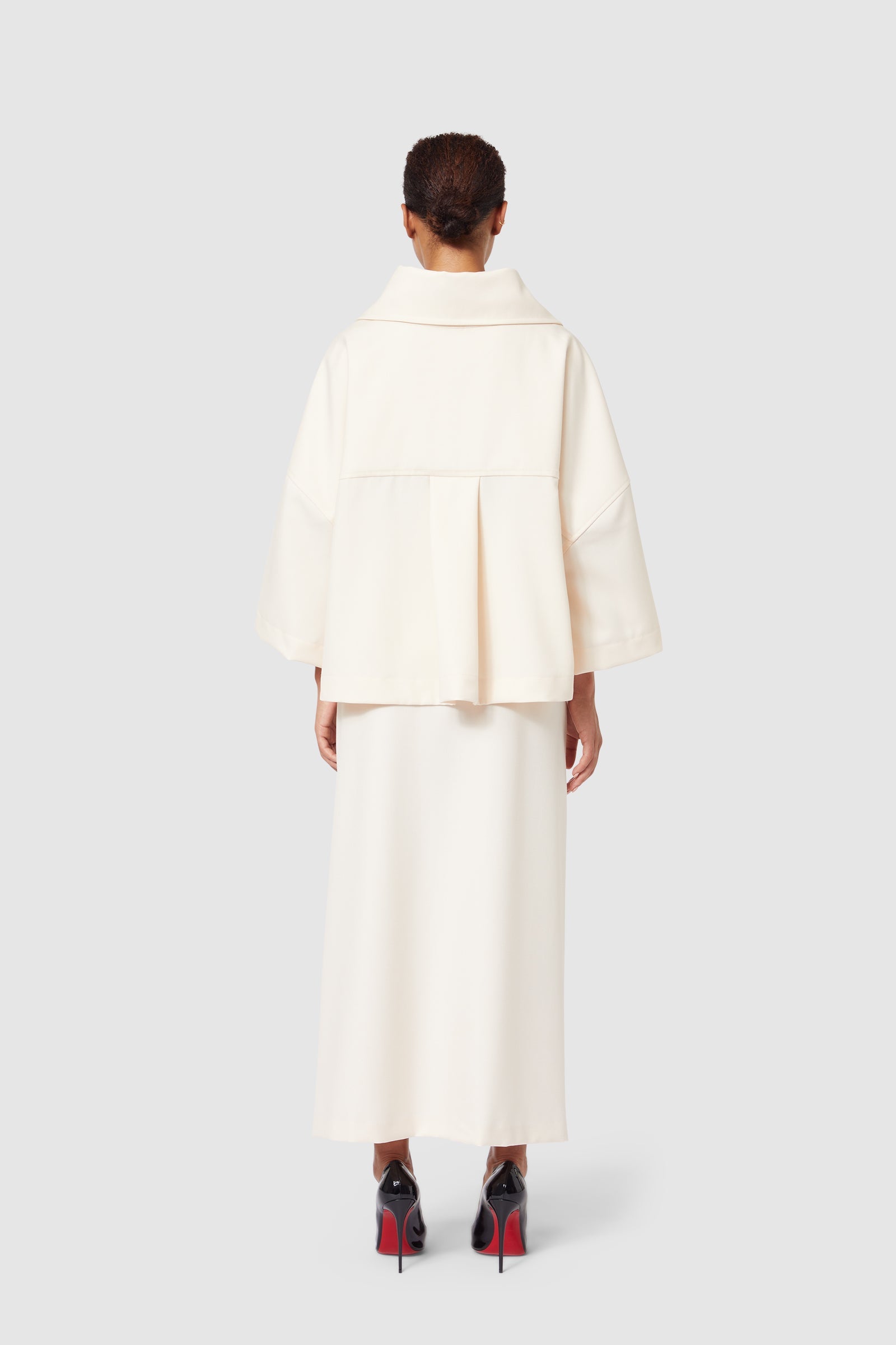 Cream oversized coat with a wide collar and pleated back, paired with matching long skirt and high-heeled shoes.