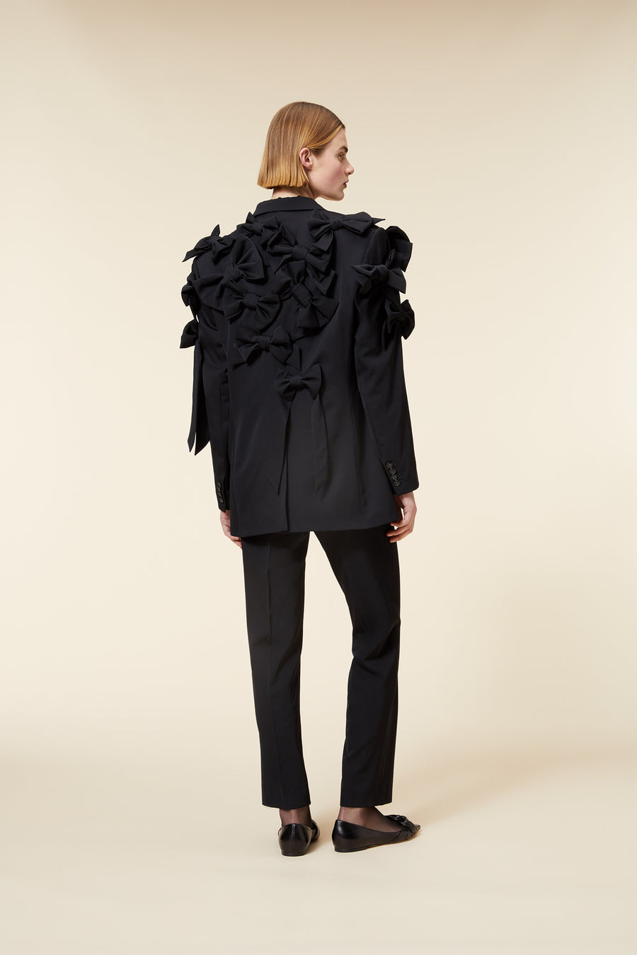 Elegant all-black suit with ruffled bow detailing, featuring tailored pants and paired with black loafers on beige background.