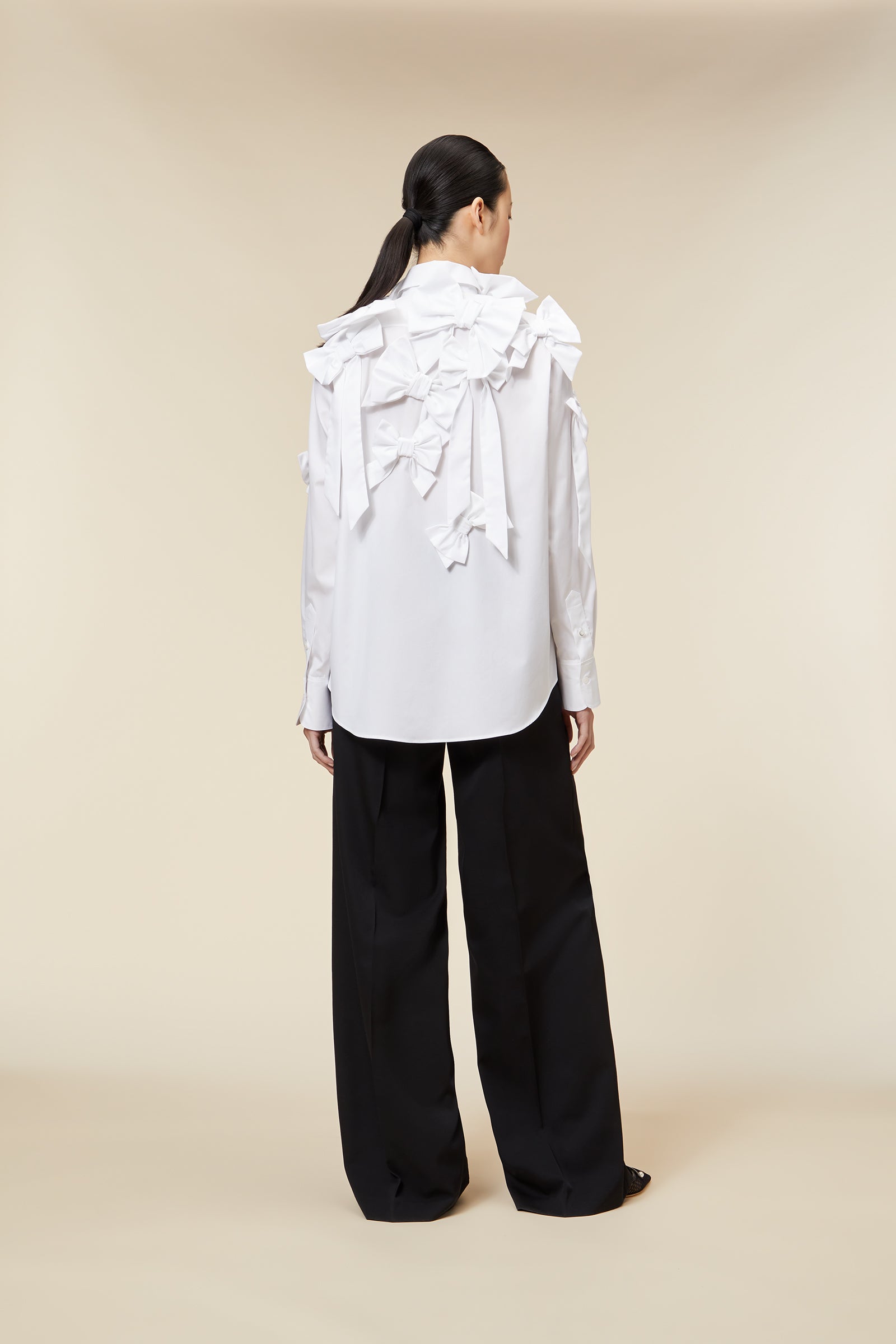 Model wearing a white ruffled blouse with bow details paired with black wide-leg trousers on a beige background.