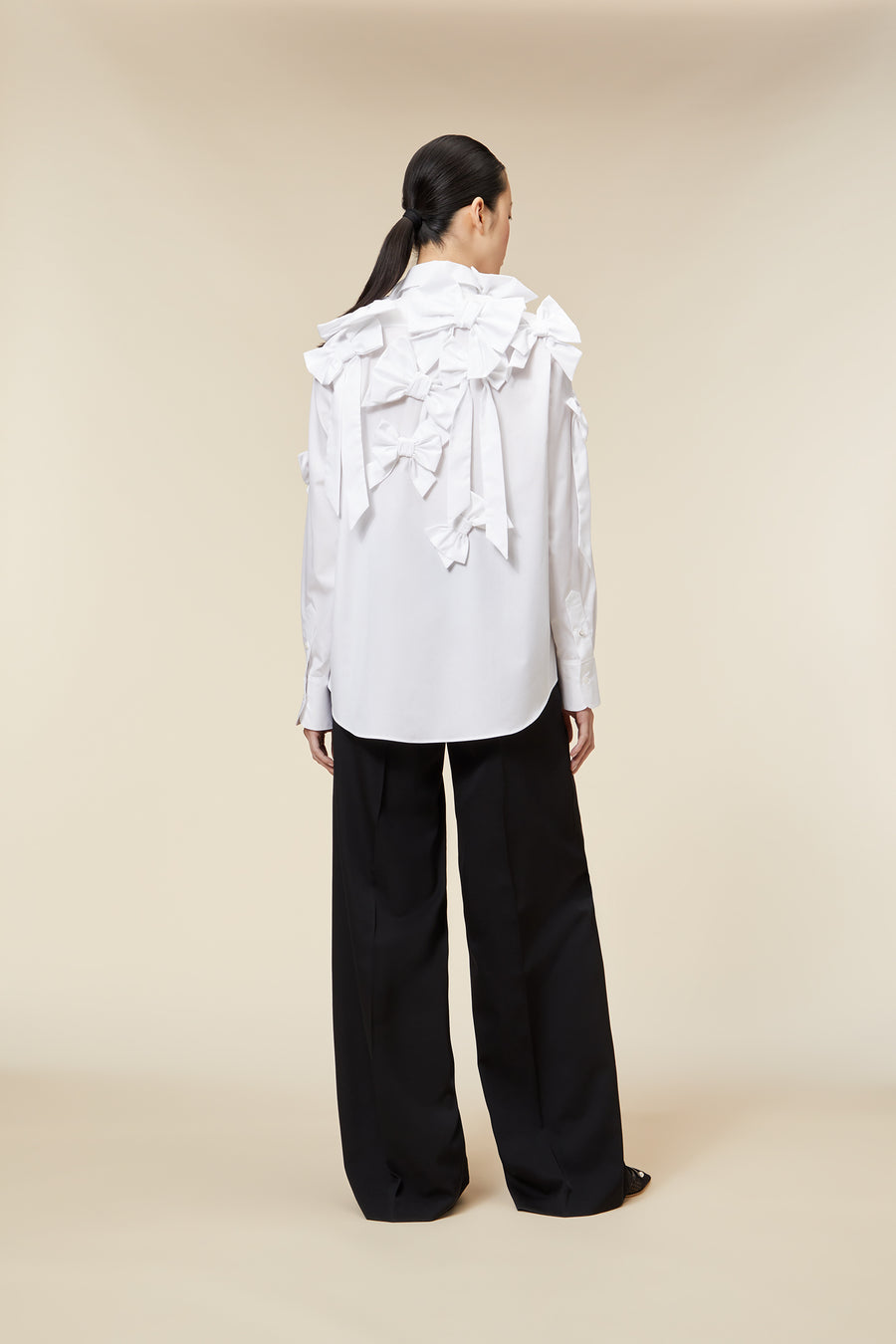 Model wearing a white ruffled blouse with bow details paired with black wide-leg trousers on a beige background.