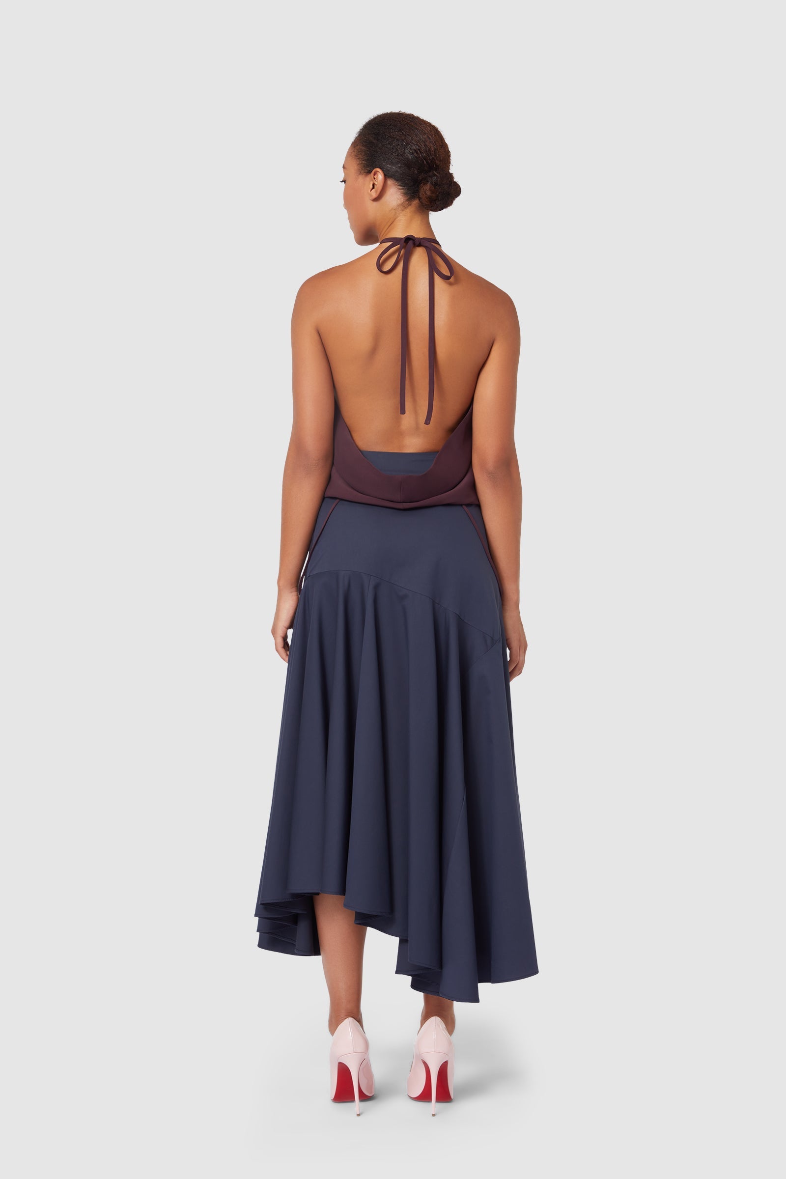 Back view of a model wearing a sleeveless halter top with an open back and an asymmetrical navy skirt paired with beige high heels.