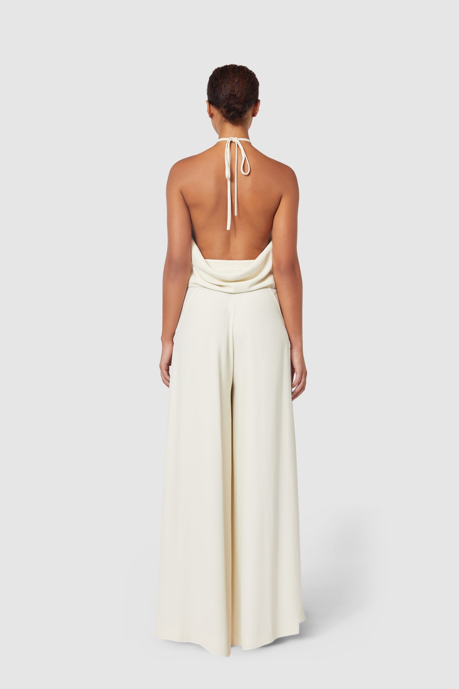 Backless ivory halter jumpsuit with draped cowl neckline, featuring wide-leg high-waisted pants and soft flowing fabric.