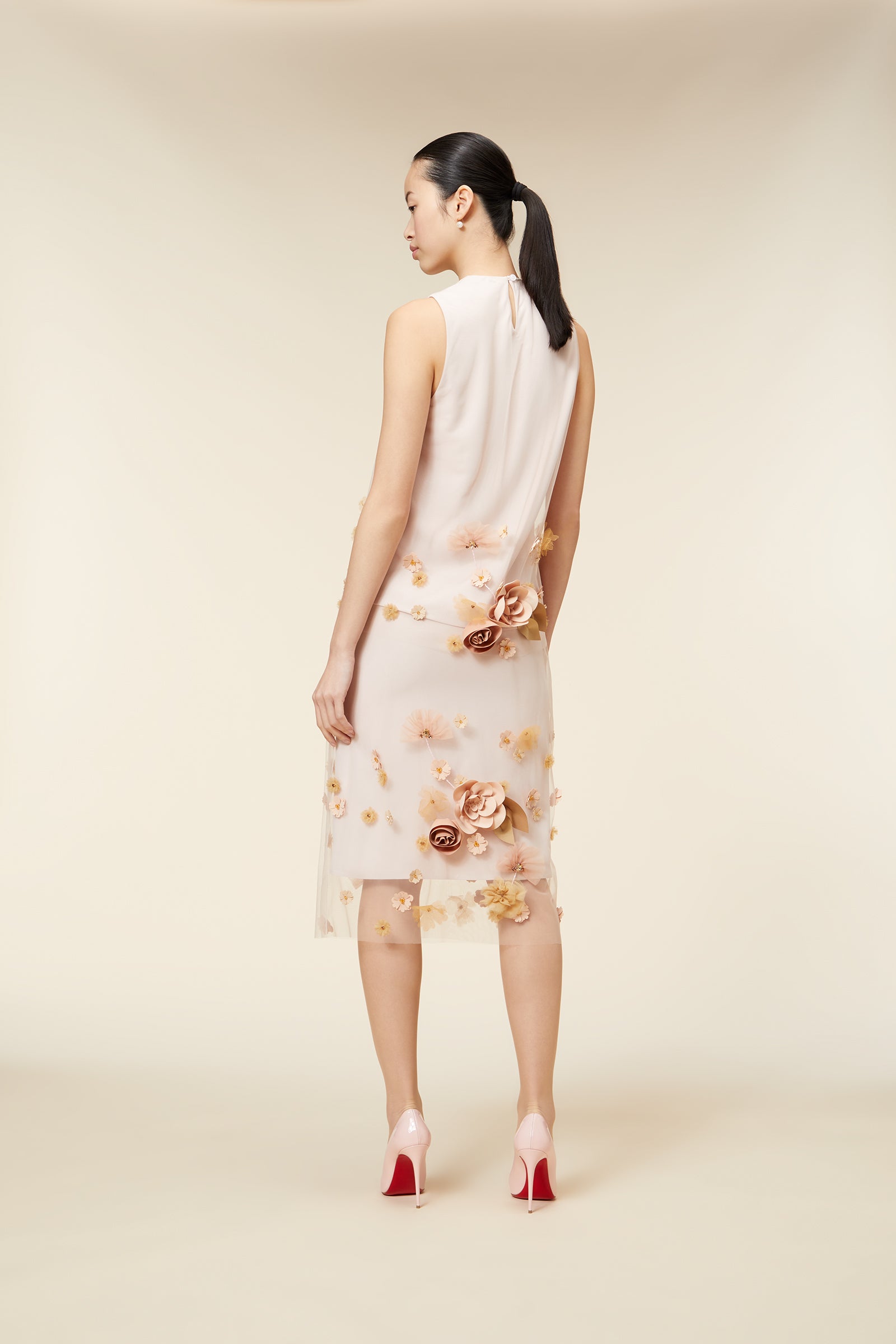 Woman wearing a sleeveless floral dress and pink high heels, standing against a neutral background, showcasing elegant style.