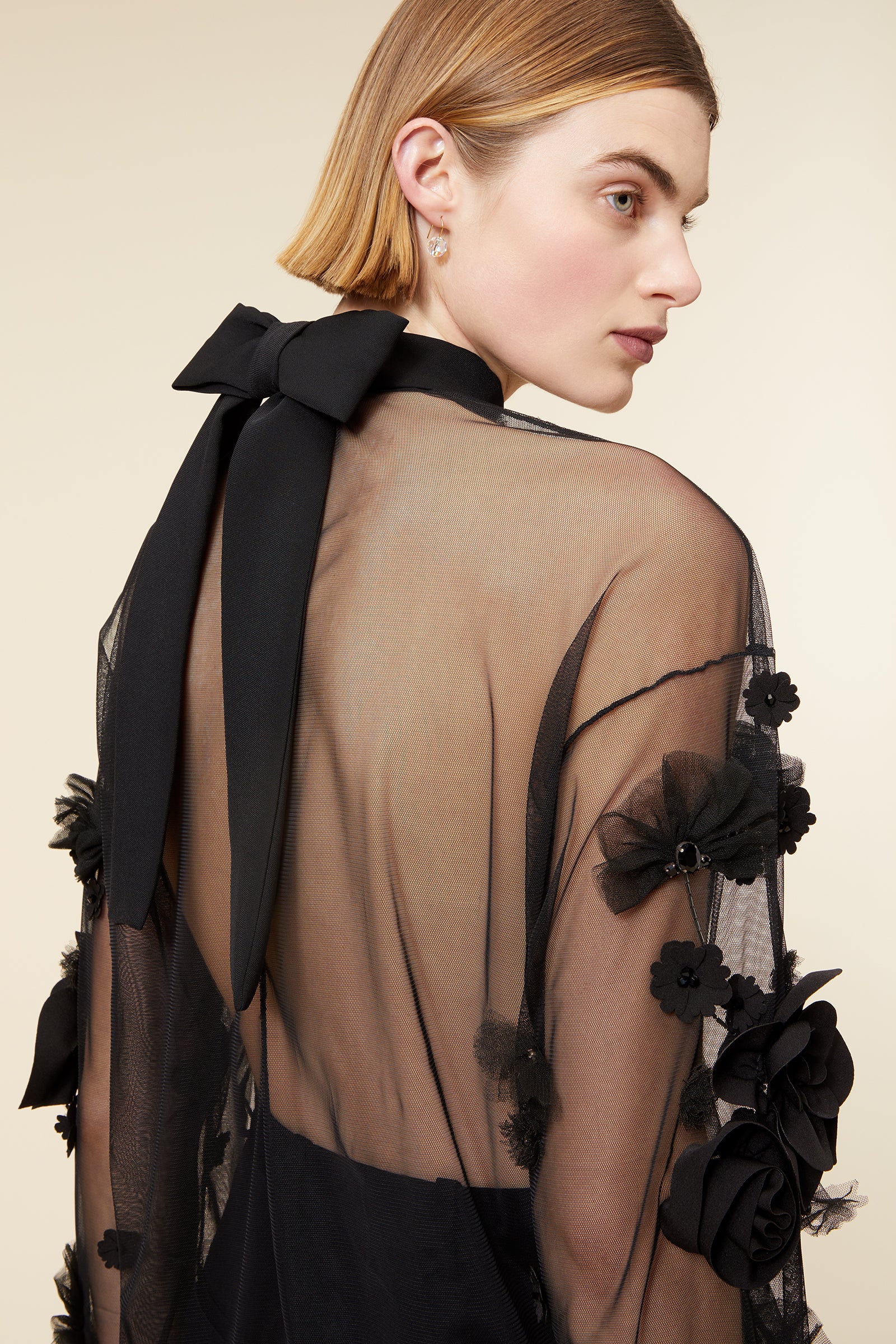 Model wearing a black sheer blouse with floral appliqué and matching trousers, posing against a neutral background.