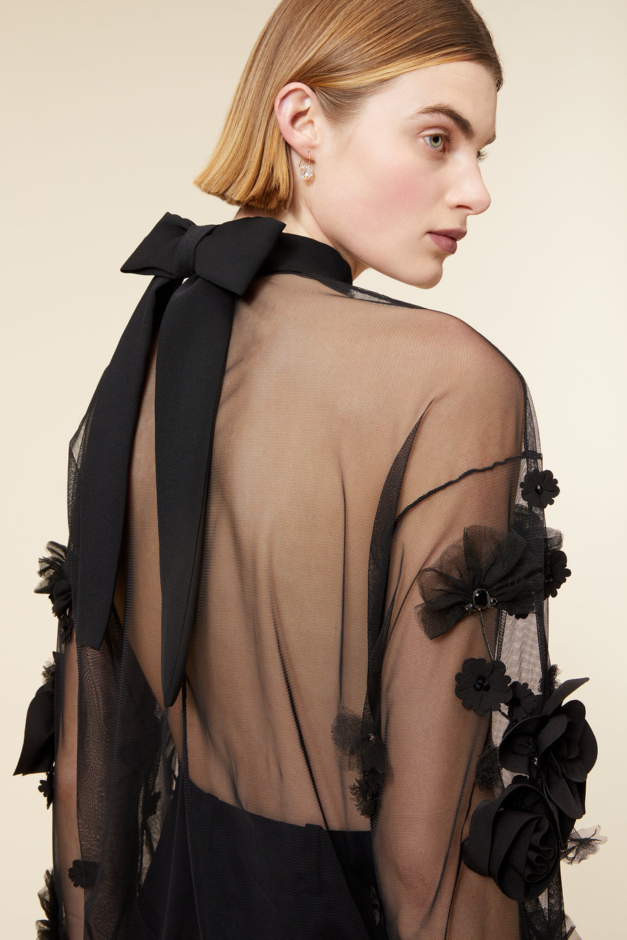 Model wearing a black sheer blouse with floral appliqué and matching trousers, posing against a neutral background.