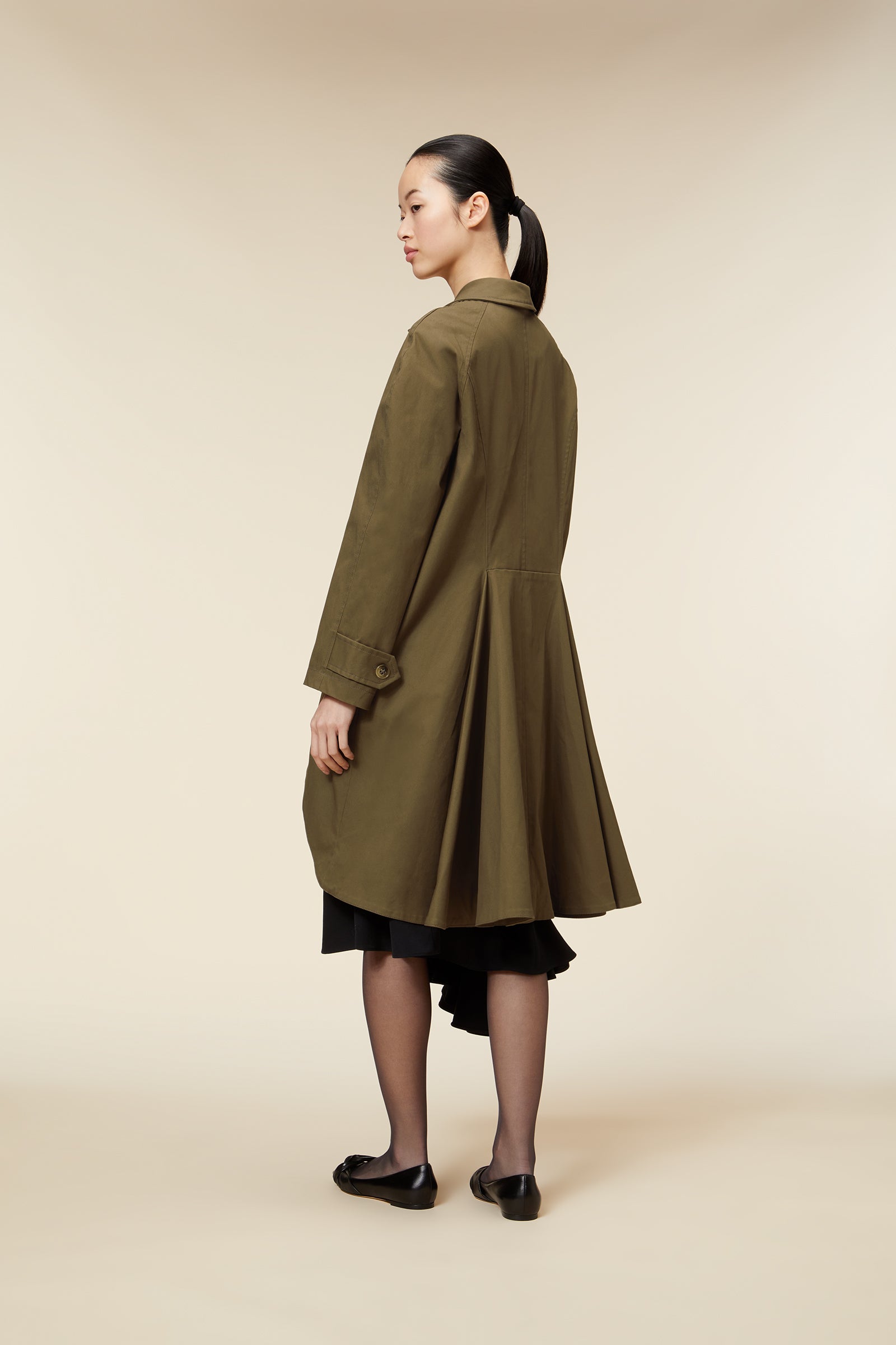 Elegant khaki trench coat features a tailored silhouette, buttoned cuffs, and a flaring hem, worn over a layered black skirt.