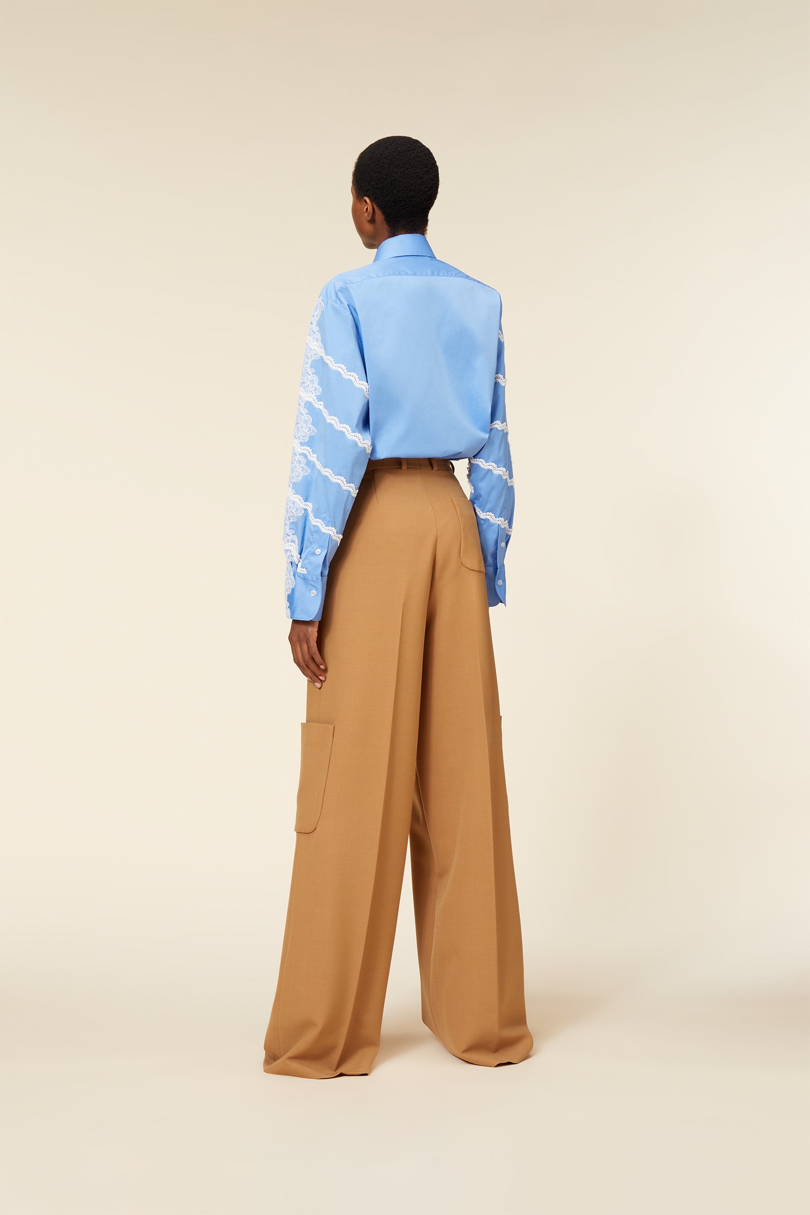 Back view of a model wearing a sky blue shirt with lace detailing and wide-legged tan trousers featuring patch pockets.