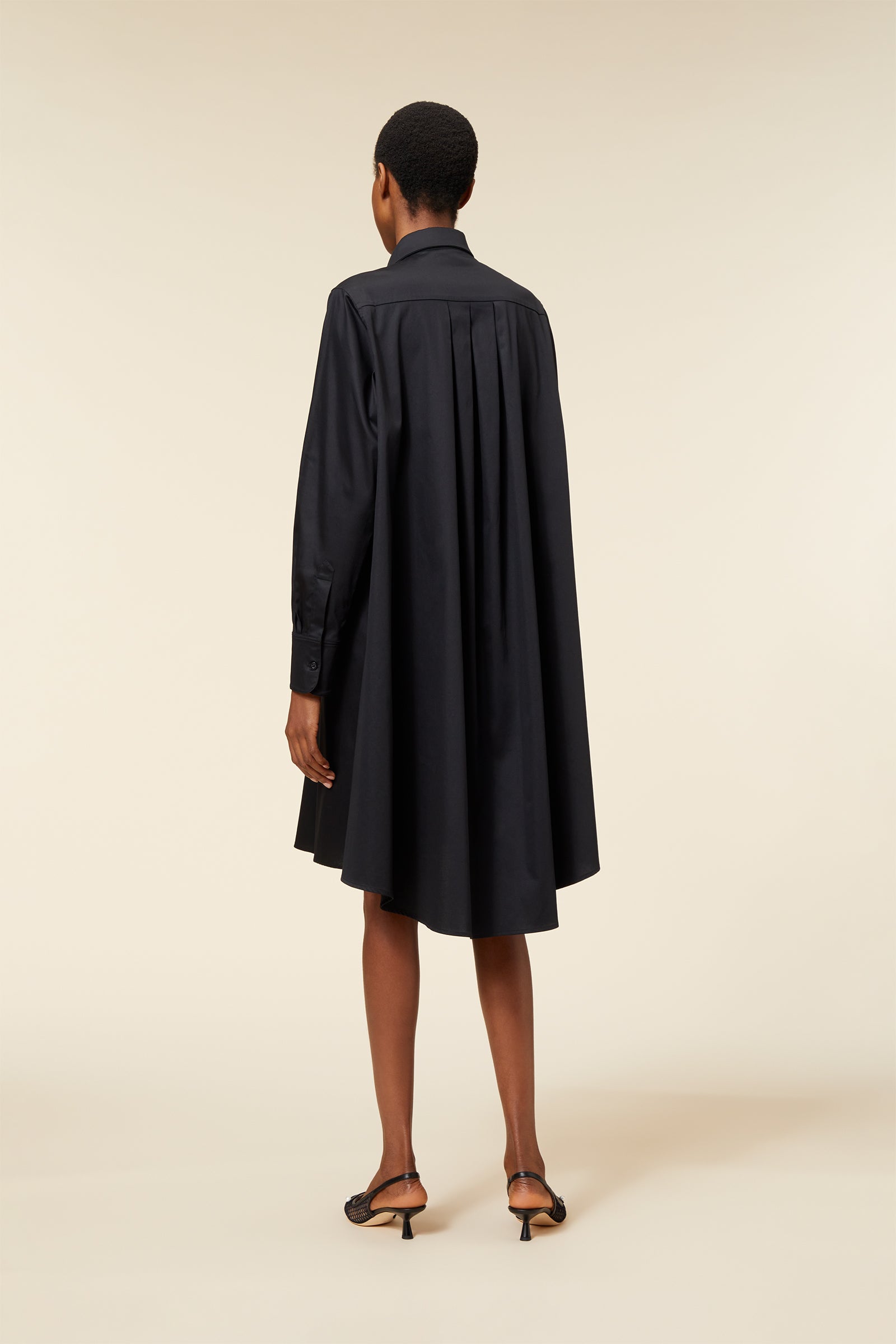 Black pleated long-sleeve shirt dress with a flowing silhouette, paired with elegant black heeled sandals.