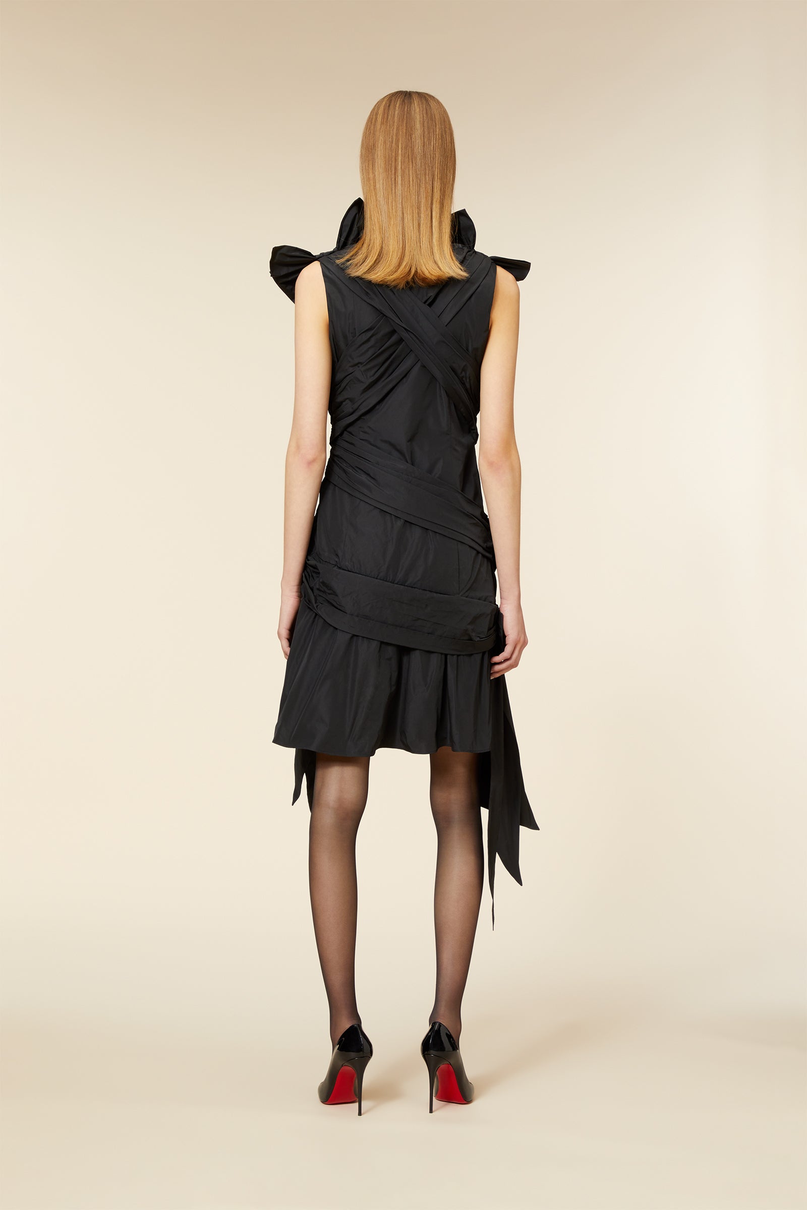 Rear view of model in elegant black knee-length dress with ruched details, paired with sheer tights and high-heeled shoes.