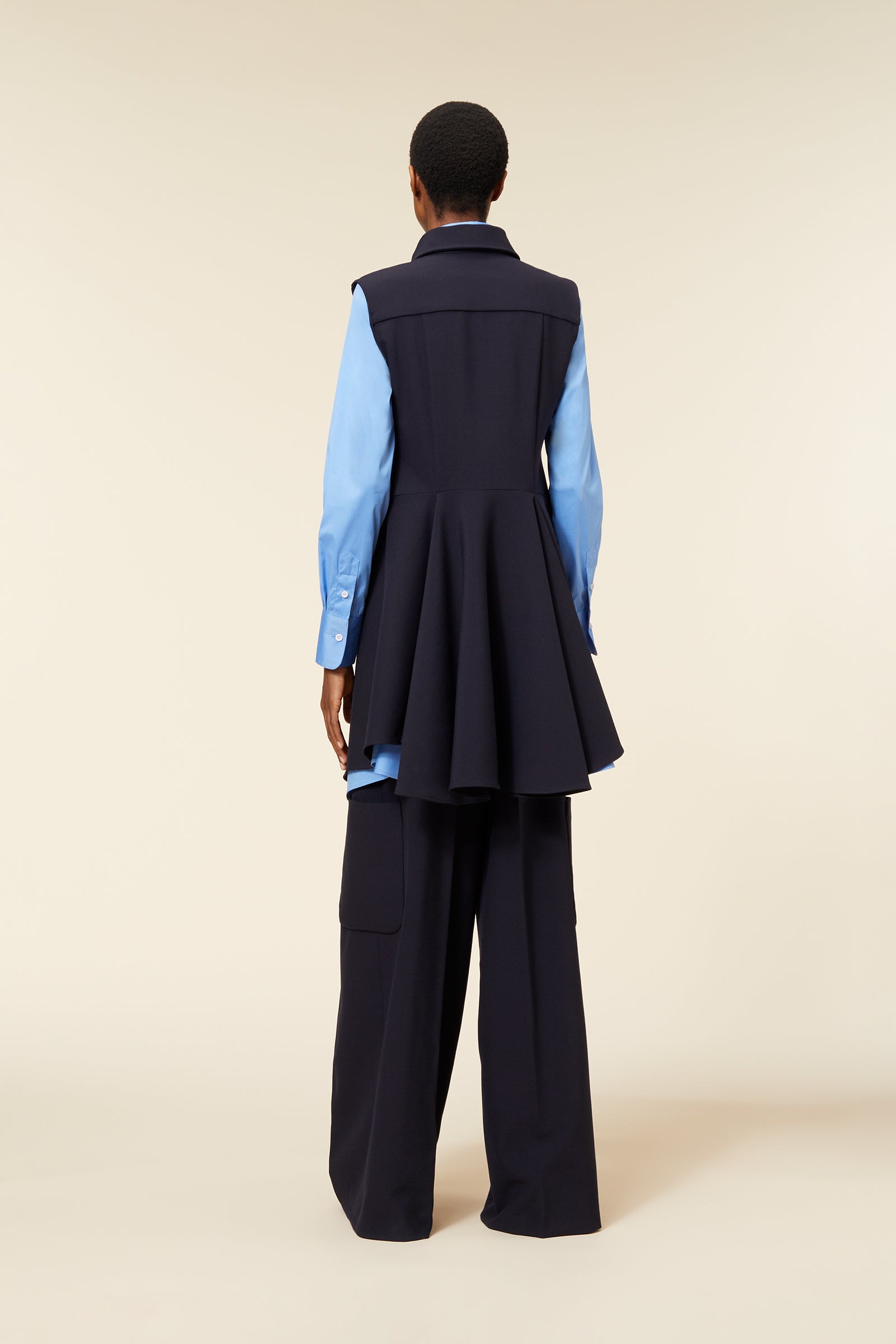 Long navy sleeveless dress with flared hem over a light blue button-up shirt, paired with wide-legged black trousers.