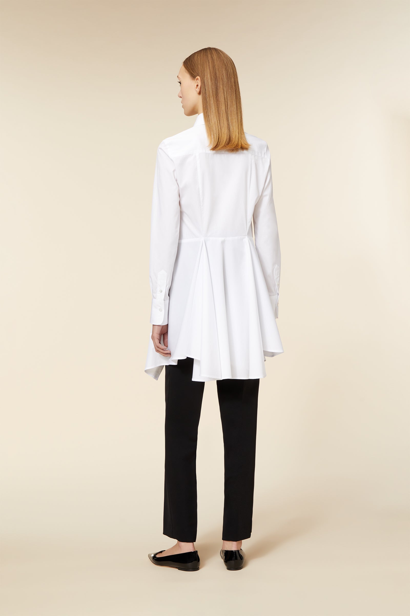 White tailored shirt with an asymmetric hem and pleats, paired with sleek black trousers for a modern minimalist look.