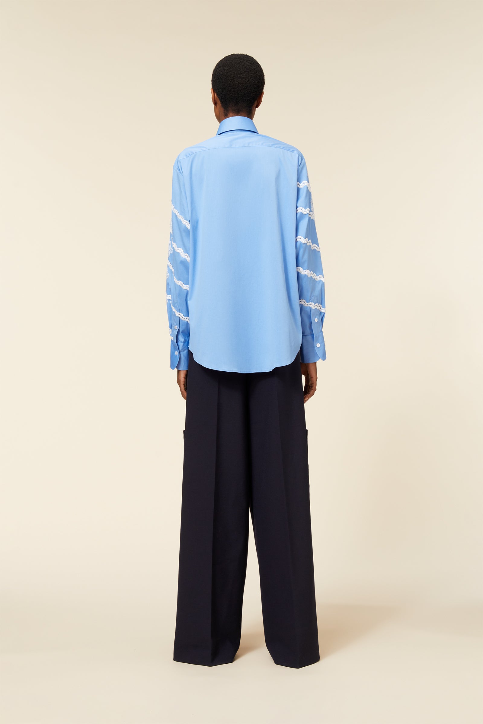 Back view of light blue button-up shirt with intricate white embroidery on sleeves, paired with navy wide-leg trousers.