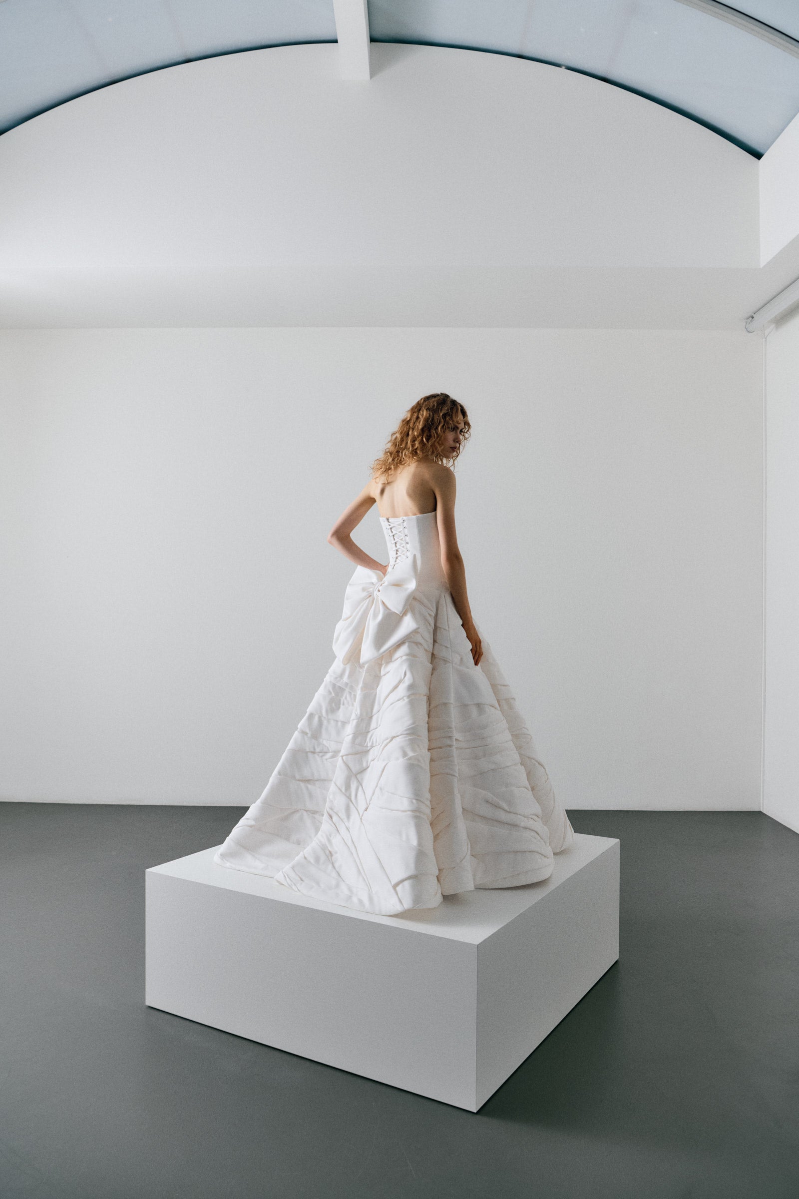 Woman in a white strapless gown standing on a white platform against a plain white wall.