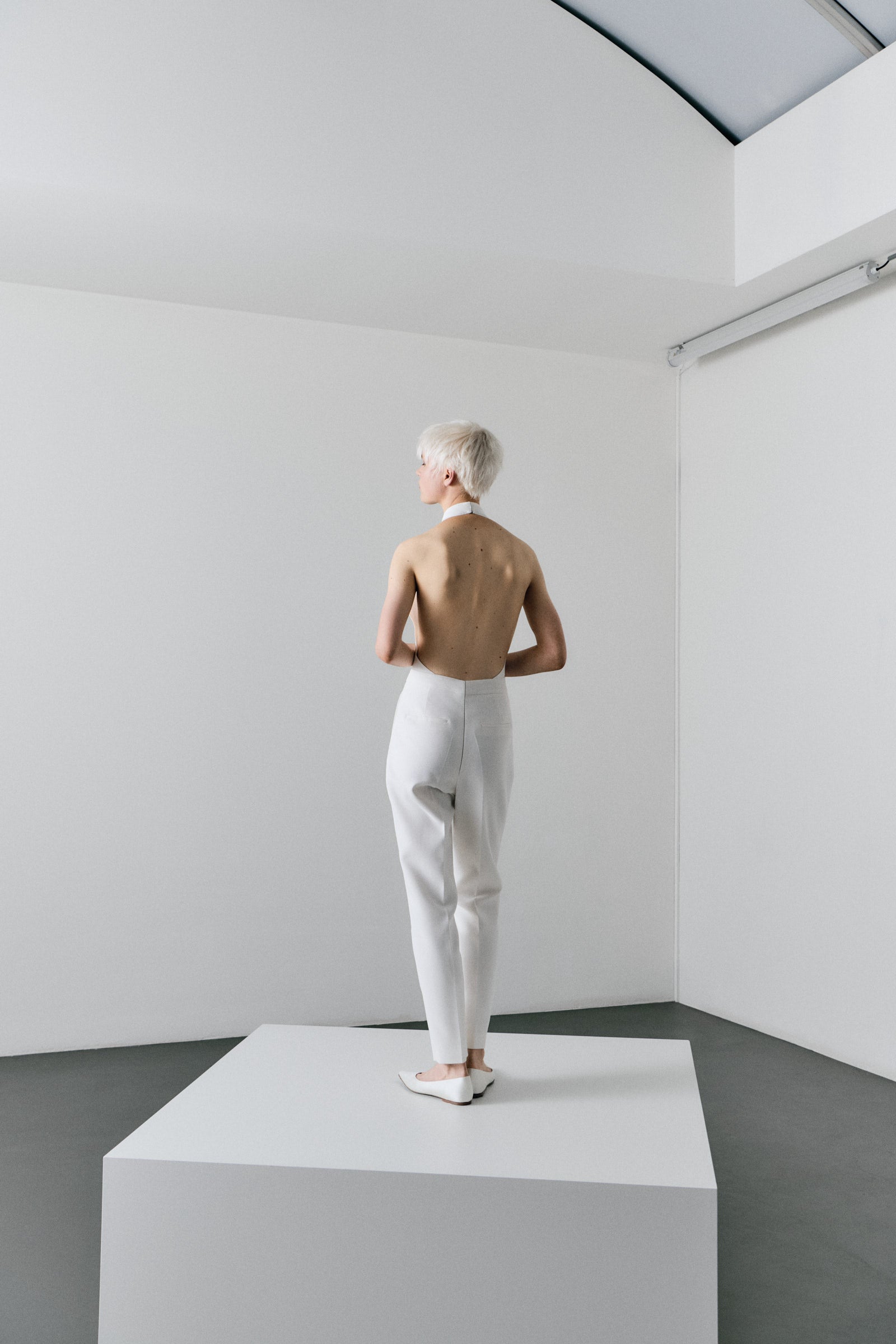 Person wearing a white outfit standing on a platform in a minimalistic room.
