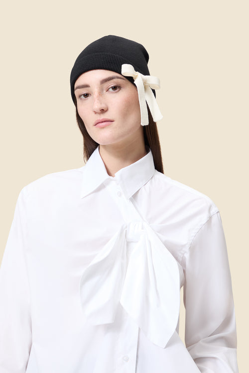 White cotton shirt with oversized bow detail paired with a black knit beanie adorned with a cream bow accent.