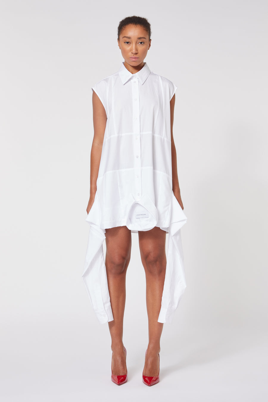 Sleeveless white shirt dress with an asymmetrical hem and button-up front, paired with vibrant red high-heeled shoes.