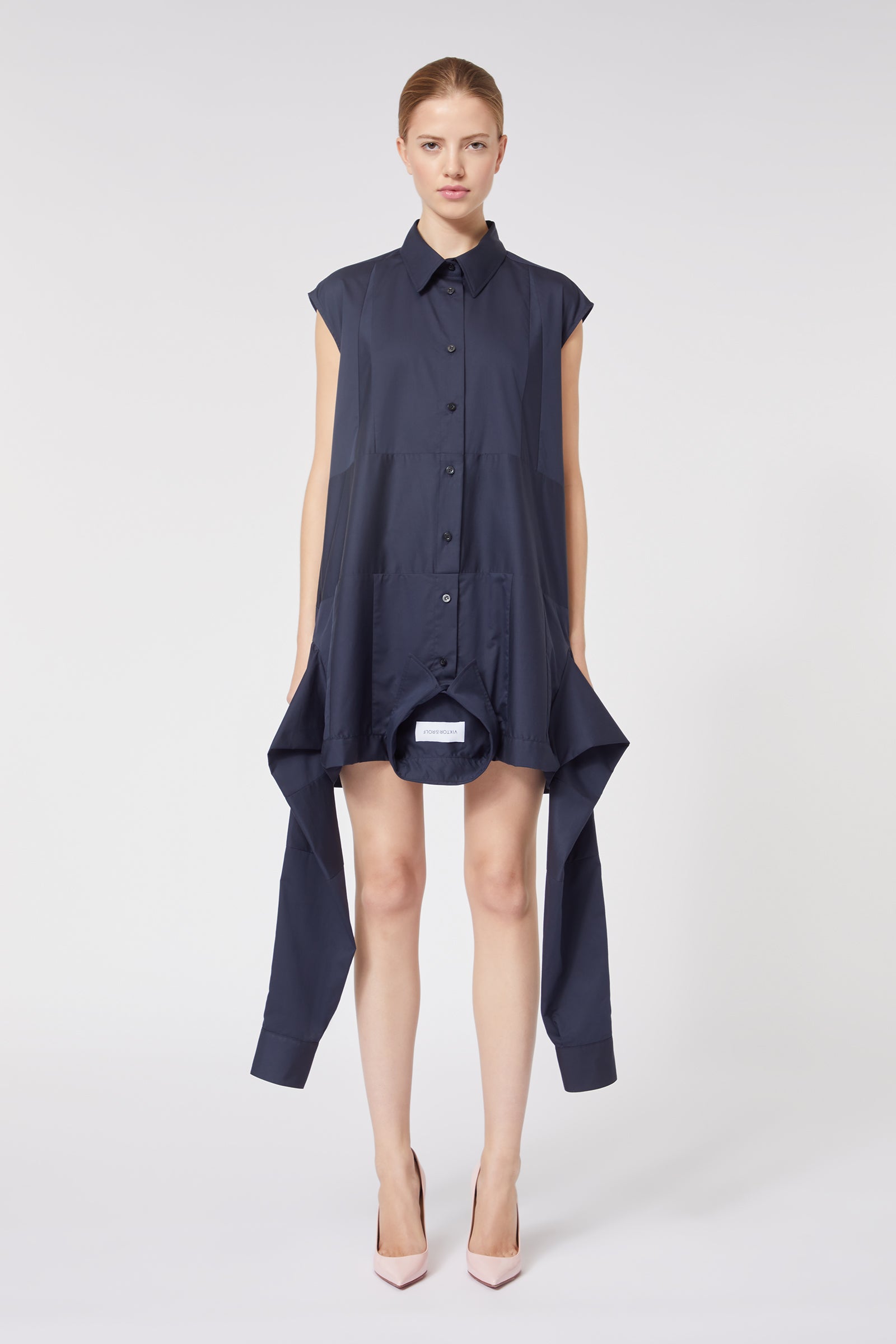 Navy blue asymmetric shirt dress with cap sleeves, buttoned front, and draped hemline, paired with pale pink heels.