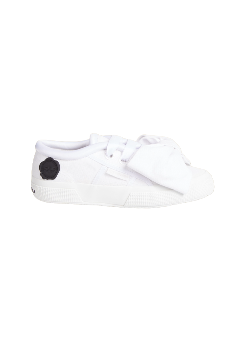 White canvas sneaker with oversized bow detailing and subtle black logo embellishment, featuring textured rubber sole.