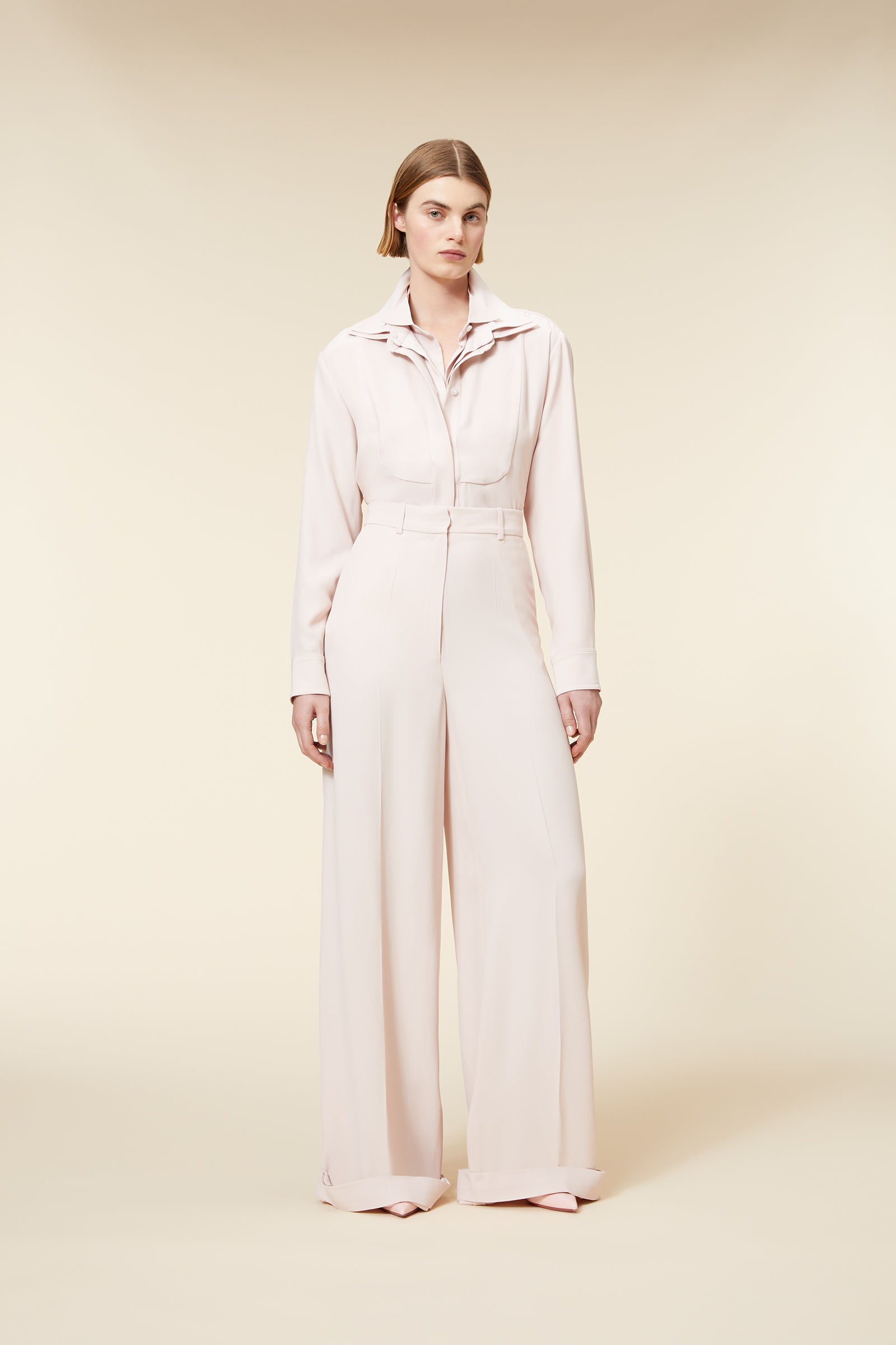 Woman wearing a light beige jumpsuit with wide-leg pants, collared neckline, and button-up front on a neutral background.