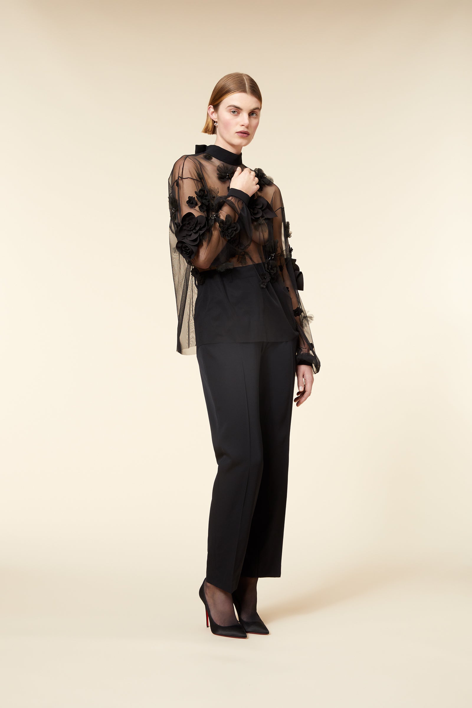 Model wearing a black sheer blouse with floral appliqué and matching trousers, posing against a neutral background.