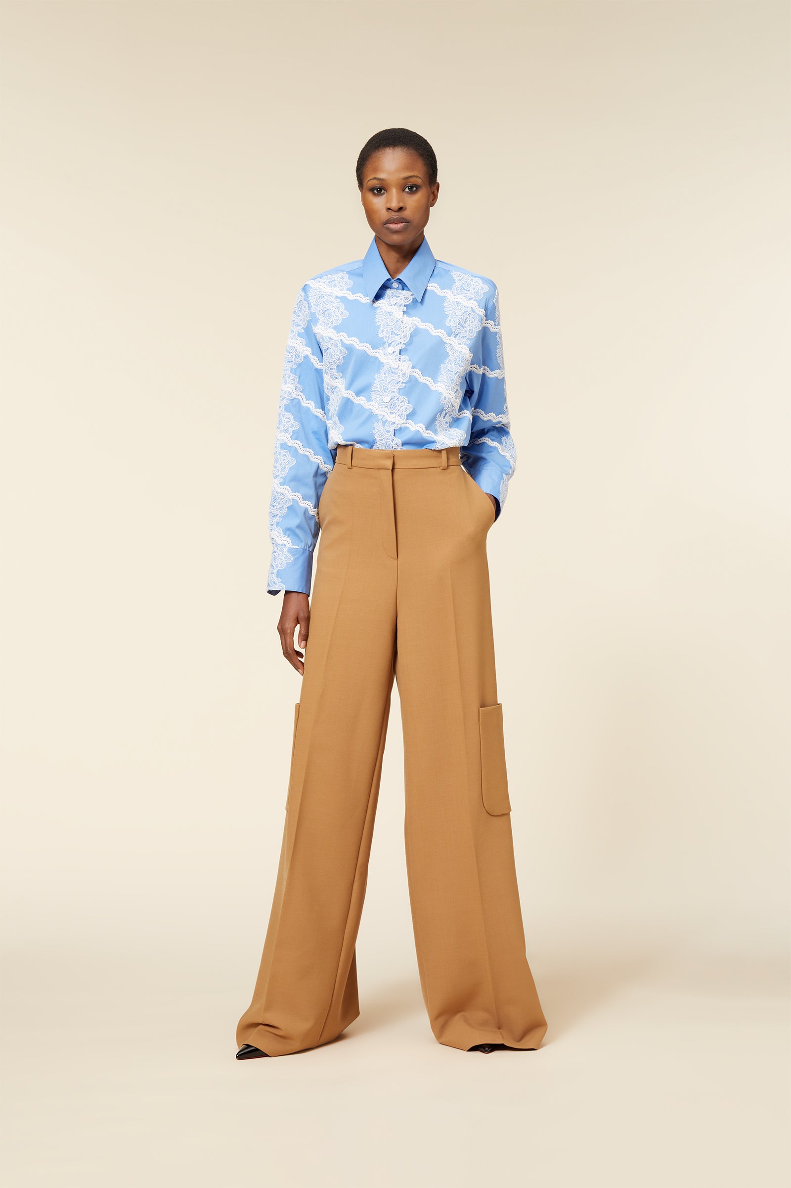 A model wears a blue cotton blouse with white lace patterns paired with high-waisted camel wool wide-leg trousers.