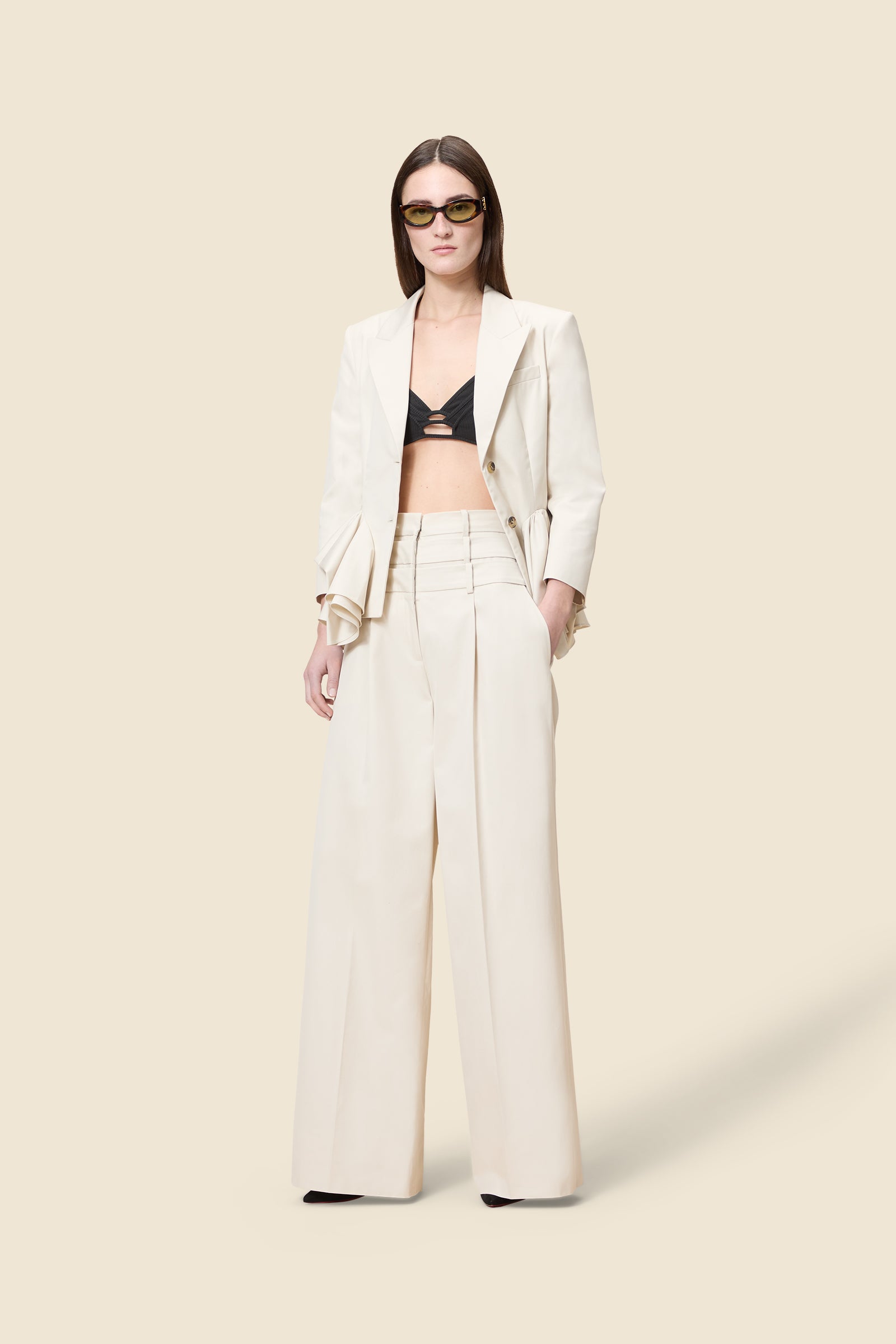 Model wearing a beige blazer with ruffles at the bottom, styled with black bralette top and high waist multi-layer beige trousers, standing on a beige background.