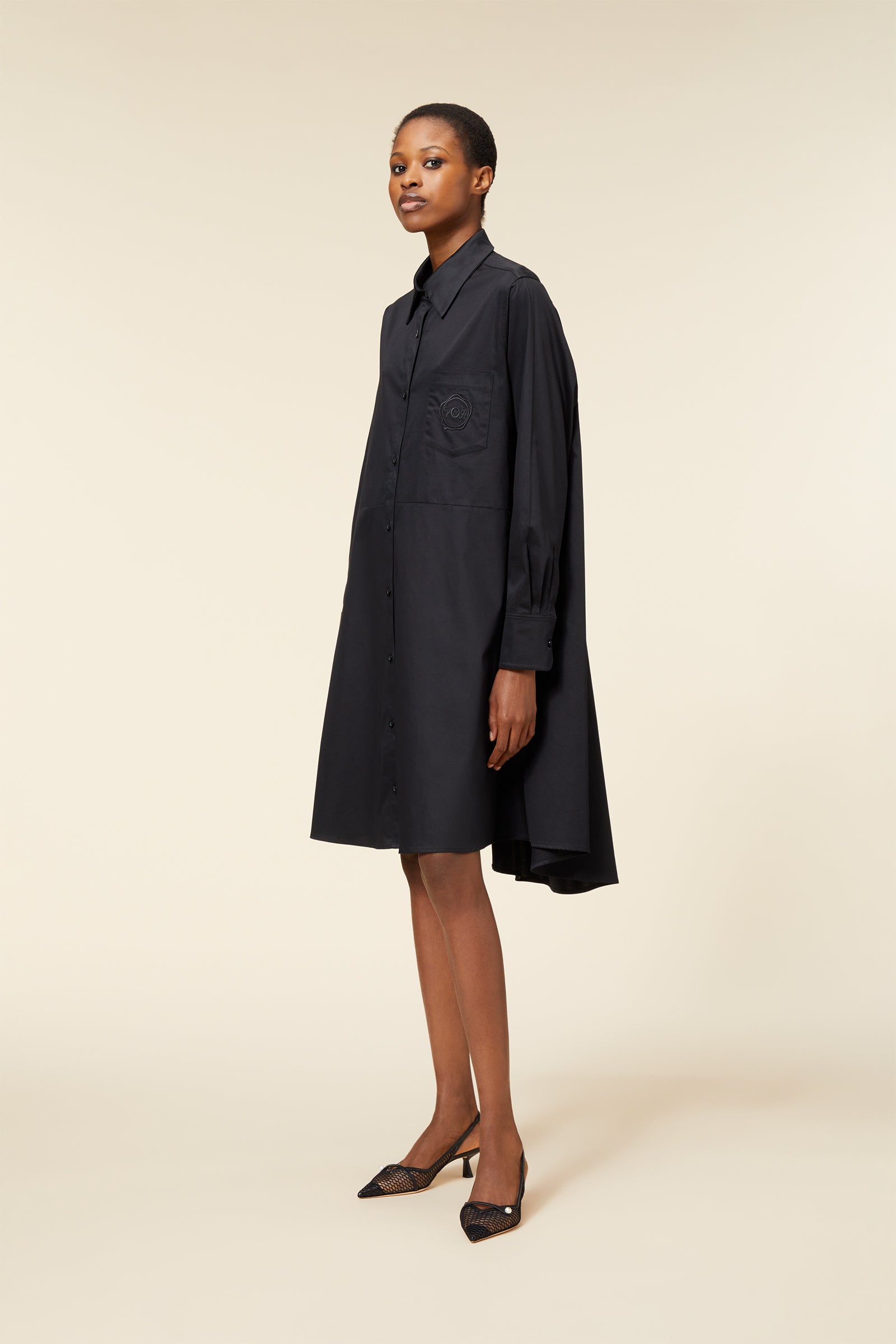Black button-down shirt dress with long sleeves and oversized fit, featuring an embroidered chest pocket, worn with mesh slingback heels.