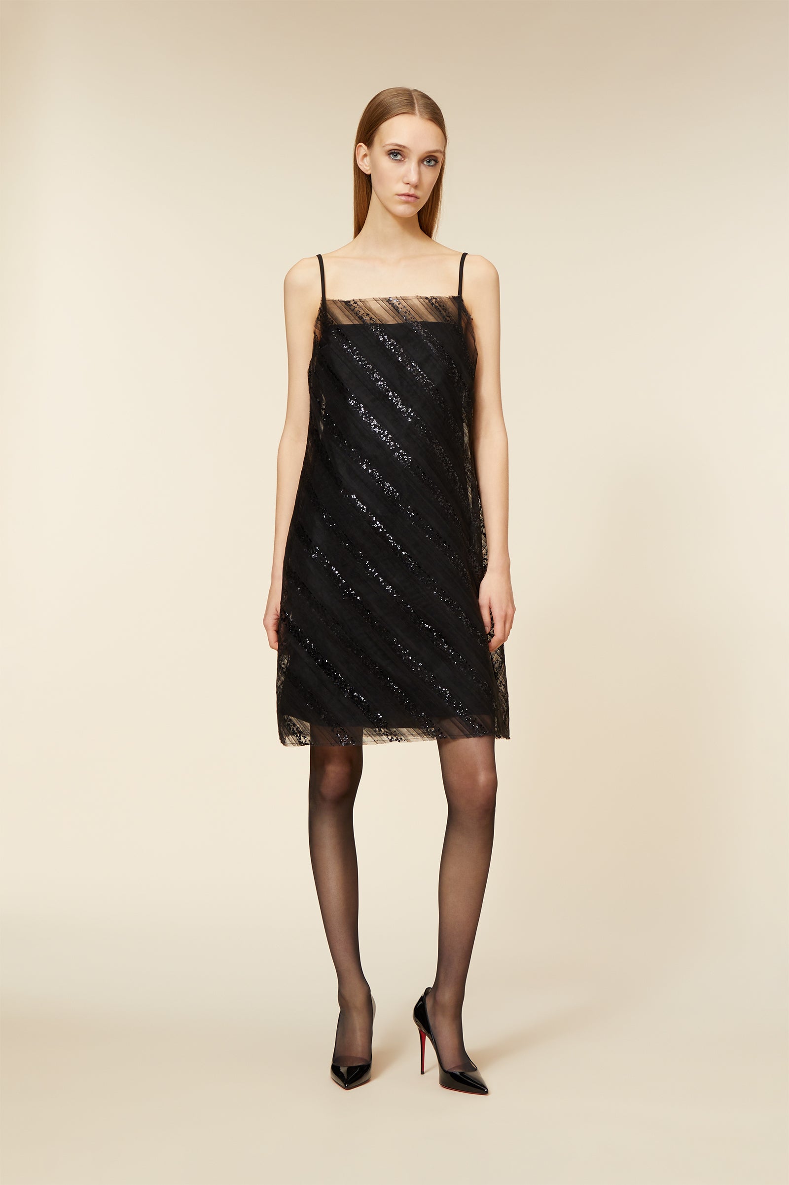 Elegant black cocktail dress with spaghetti straps and sheer overlay, paired with sheer black stockings and sleek black heels.