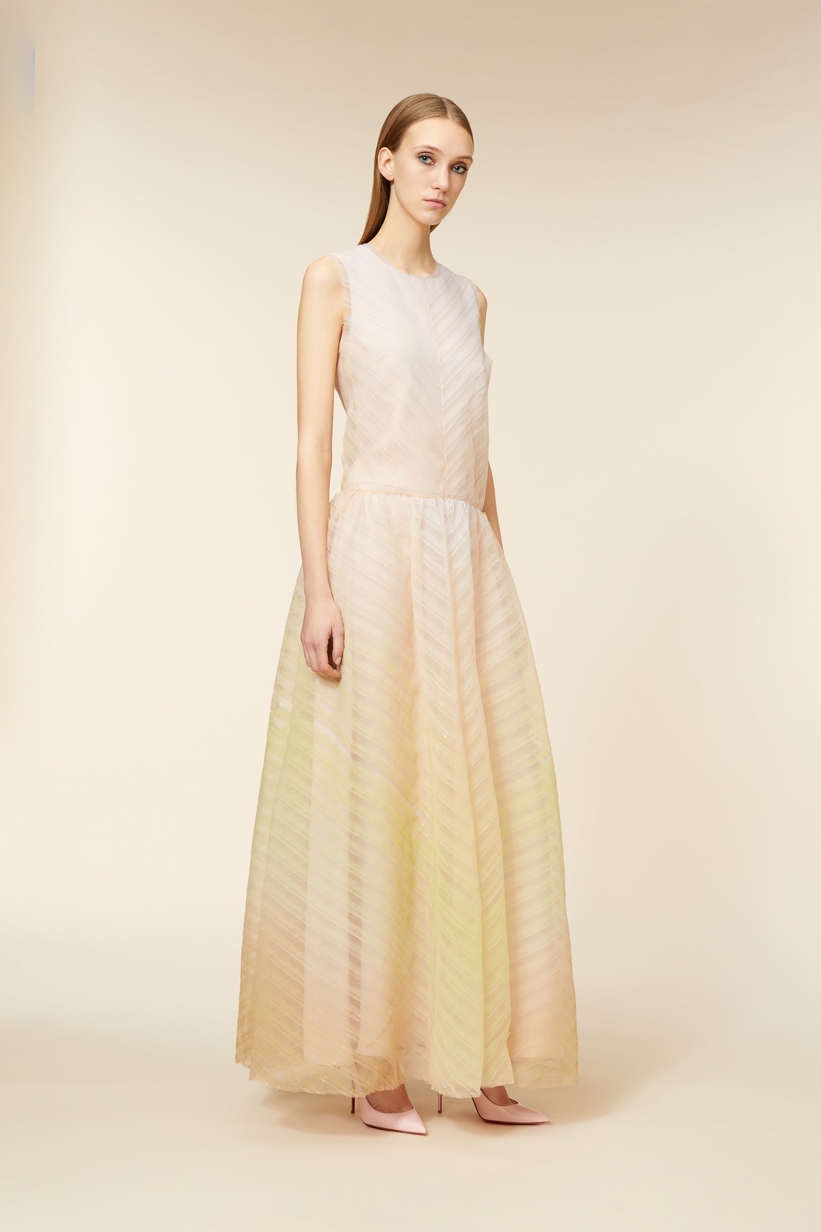 Elegant woman in a sleeveless, sheer pastel gown with diagonal stripes standing in a minimalist, soft-toned setting.