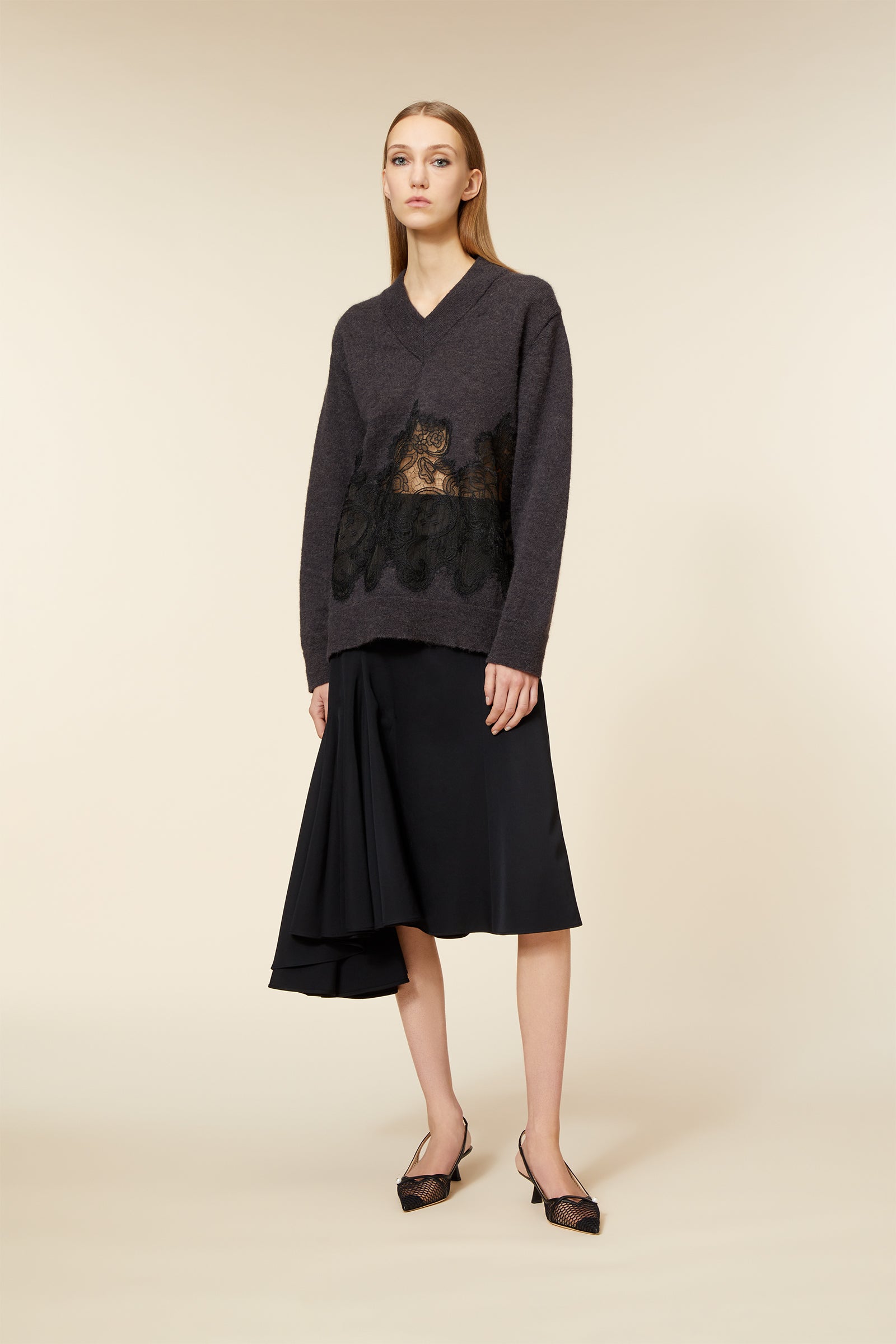 Model wearing a charcoal knit sweater featuring black lace detailing paired with an asymmetrical black skirt and pointed heels.