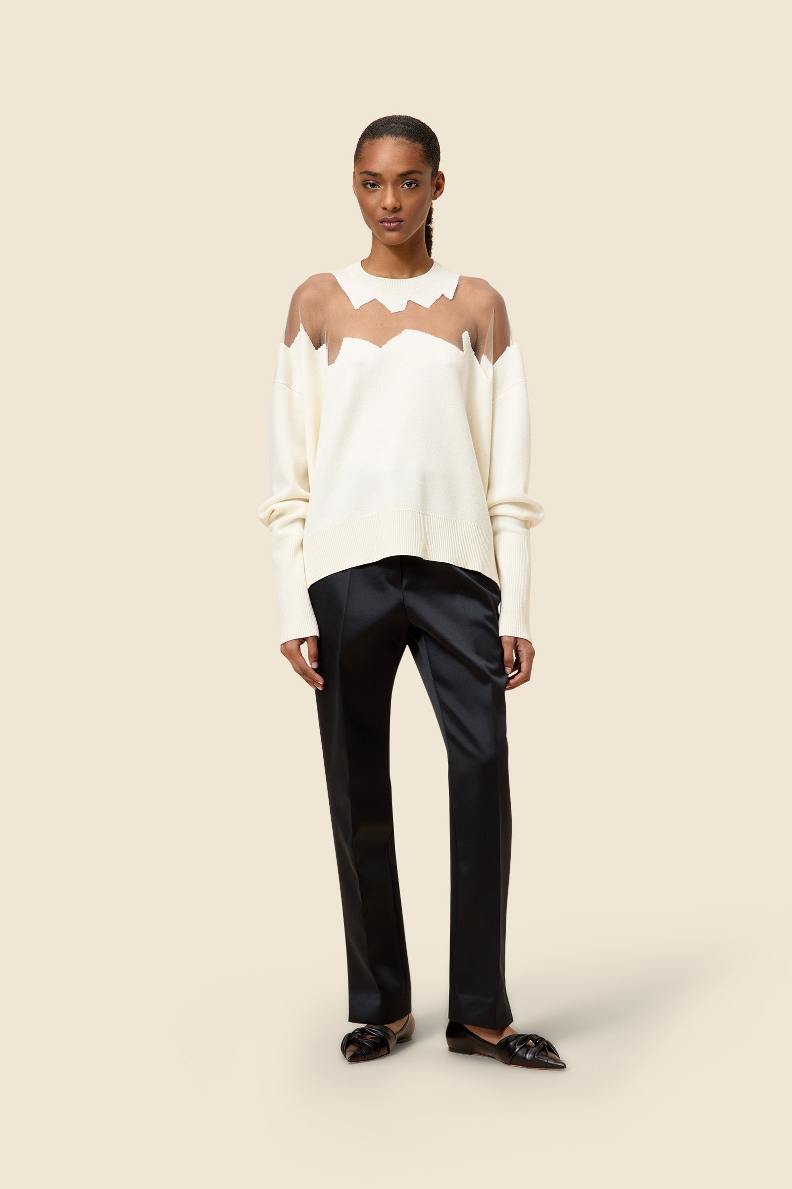 Model wearing a cream sweater with sheer zigzag design and sleek black straight-leg trousers on a plain background.