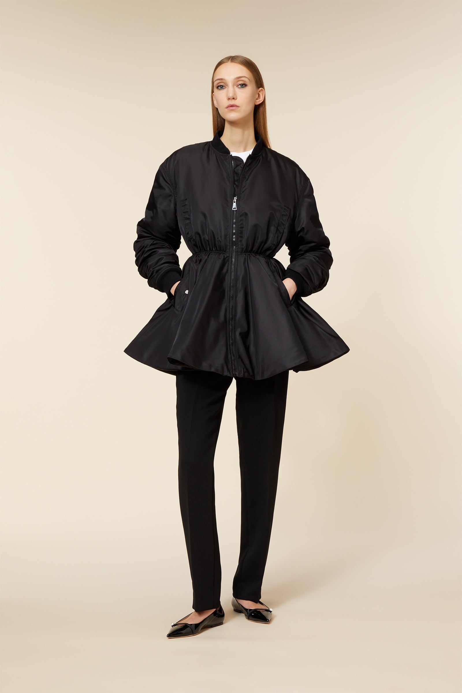 Model in a sleek black satin bomber jacket with a cinched waist, paired with tailored black trousers and pointed flats.