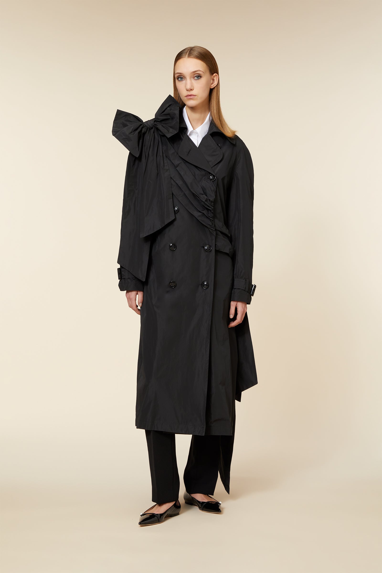 Elegant black trench coat with double-breasted buttons and large asymmetric bow detail, paired with chic black trousers.